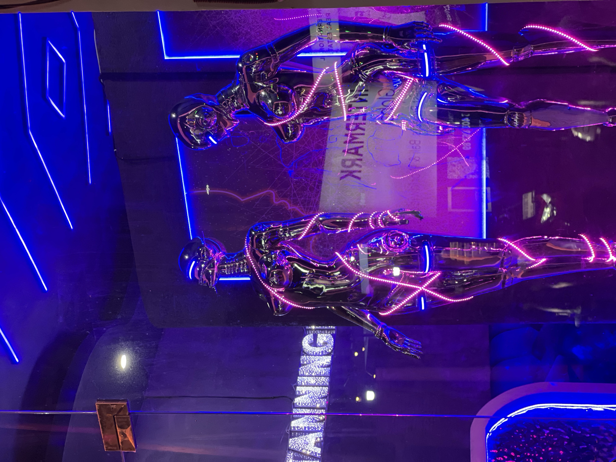 The image shows a display window, likely of a retail store in North Kuta, Indonesia, featuring two chrome-plated humanoid figures with neon pink light strands draped over their bodies. The figures are positioned one above the other, with the top figure reflected in the glass surface. A reversed text appears on the surface, potentially related to the store's name. Neon blue lighting outlines architectural elements in the background. A banknote with Indonesian text is faintly visible behind the figures.