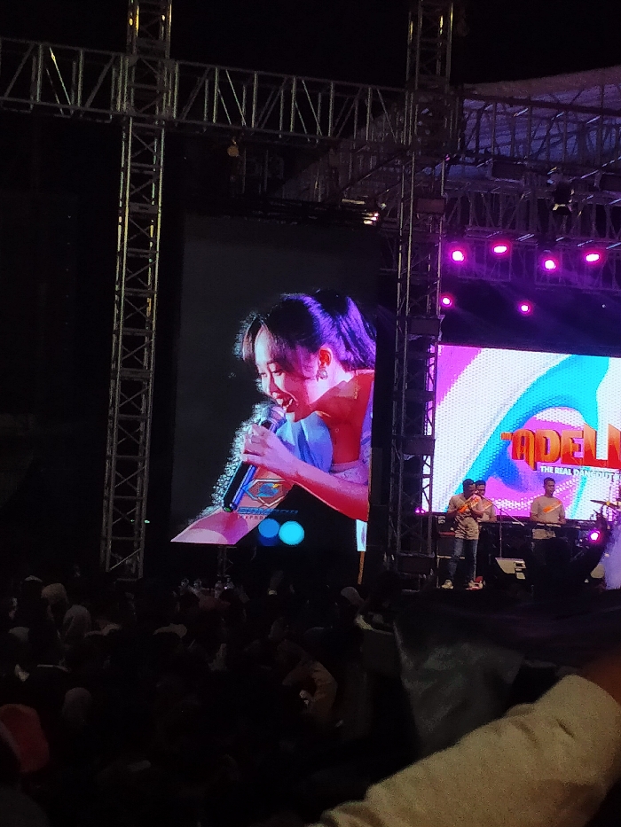 The image shows a concert stage in Boyolali, Indonesia, at the coordinates -7.535 latitude, 110.601 longitude. A large screen displays a woman singing into a microphone, holding the microphone with her right hand. She is wearing a blue top and a light pink scarf or blouse. The screen is positioned on a large metal stage with a metal scaffolding structure behind it. In the background, on the right side of the screen, there is a stage with a large screen showing a band performing with a drummer, a guitarist, and a bassist. The performance is being attended by a crowd, which is largely obscured by the screen and the foreground. The overall scene is one of a lively, energetic concert.