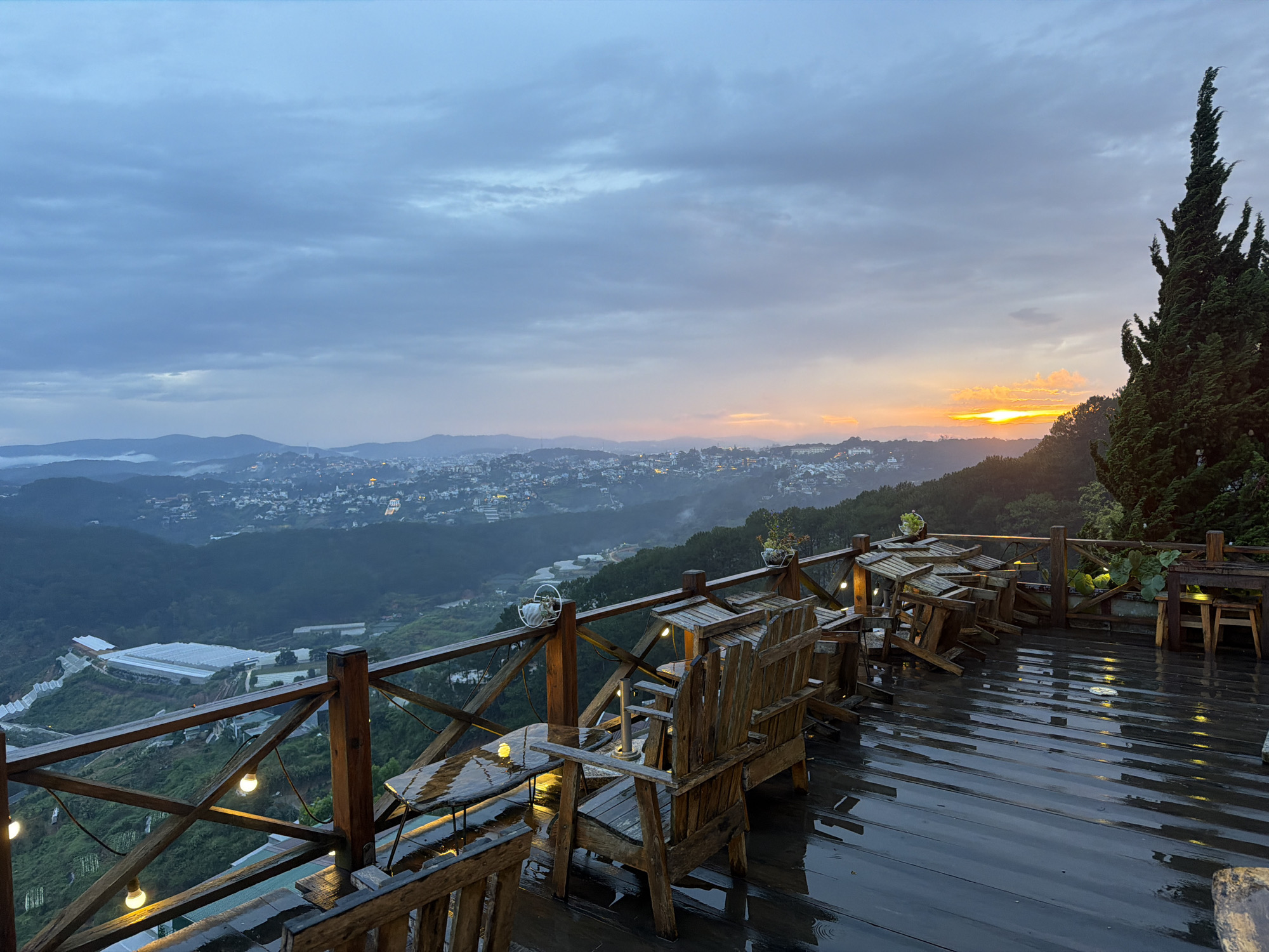An exterior, panoramic, eye-level shot captures a misty, overcast dawn over a sprawling, verdant valley from a wooden deck. The deck, wet from recent rain, reflects the dim morning light. Several rustic wooden chairs and tables are arranged on the deck, some overturned, suggesting the establishment is either closed or preparing for the day. String lights are visible, casting a warm glow on the deck. In the distance, a city unfolds across the hilly landscape, shrouded in a light fog, with the silhouette of mountains forming a backdrop. The sun is beginning to rise on the right, peeking through the clouds and casting a golden-orange hue across the horizon. A tall, dark evergreen tree stands prominently on the right side of the frame, framing the sunrise. The scene appears to be a viewpoint café or restaurant in Xuân Thành, Vietnam, offering a scenic overlook of the city and valley below.