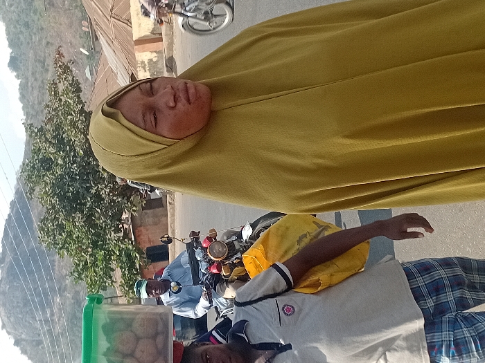 A woman wearing a yellow hijab is featured in the upper portion of the image, a motorbike with two men riding in the lower portion of the image. The background includes buildings, trees, a large hill, and overhead power lines. The men on the motorbike appear to be driving on a road. The light and perspective of the photo suggest it was taken in daylight, outside.