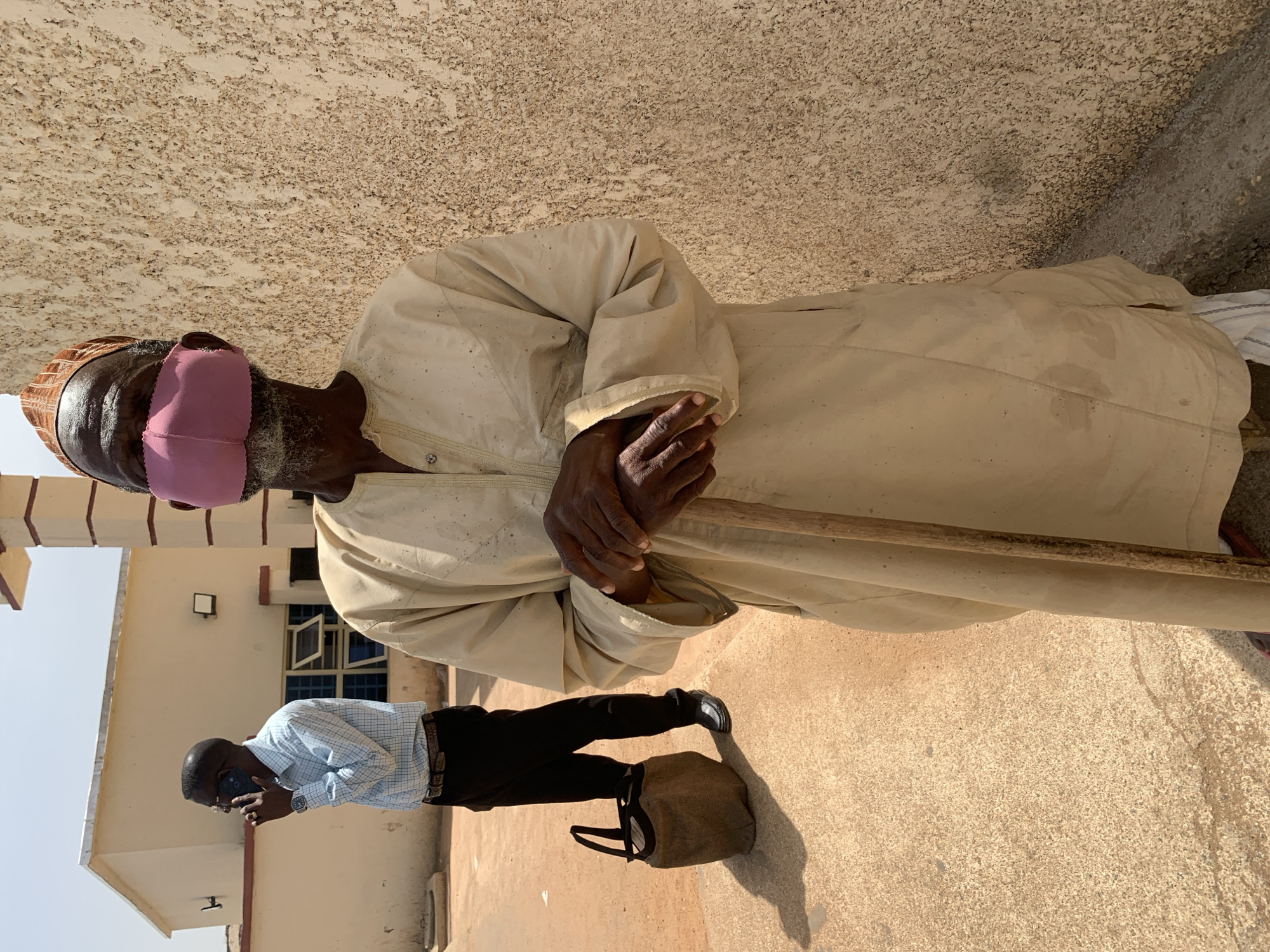 An elderly man stands with a wooden walking stick. He wears a long, light-colored robe, a woven cap, and a pink face covering. In the background, a man in a blue checkered shirt and black pants is on a cell phone next to a bag and a light-colored building with dark-framed windows. The scene appears to be set in Jos, Nigeria, suggested by the architectural style and clothing.