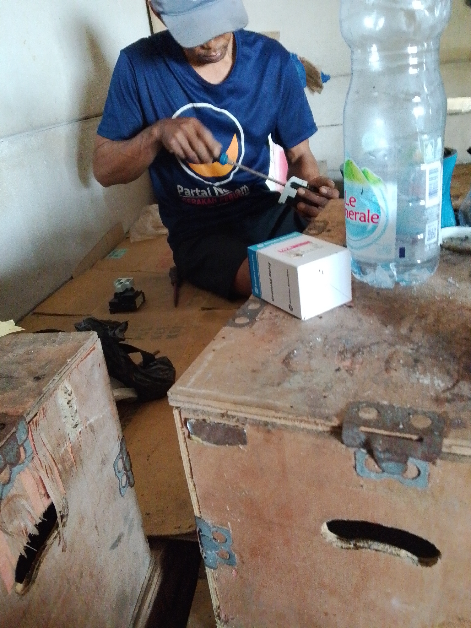 A man in Bukit Ketok, Indonesia, wearing a blue t-shirt and a gray cap, sits on a cardboard-covered floor and manipulates a metal object with a small tool. To his right is a white box labeled "Overload Relay" and a plastic water bottle. An electrical device and a dark-colored bag are nearby on the floor. In the foreground are two wooden boxes with metal hardware.