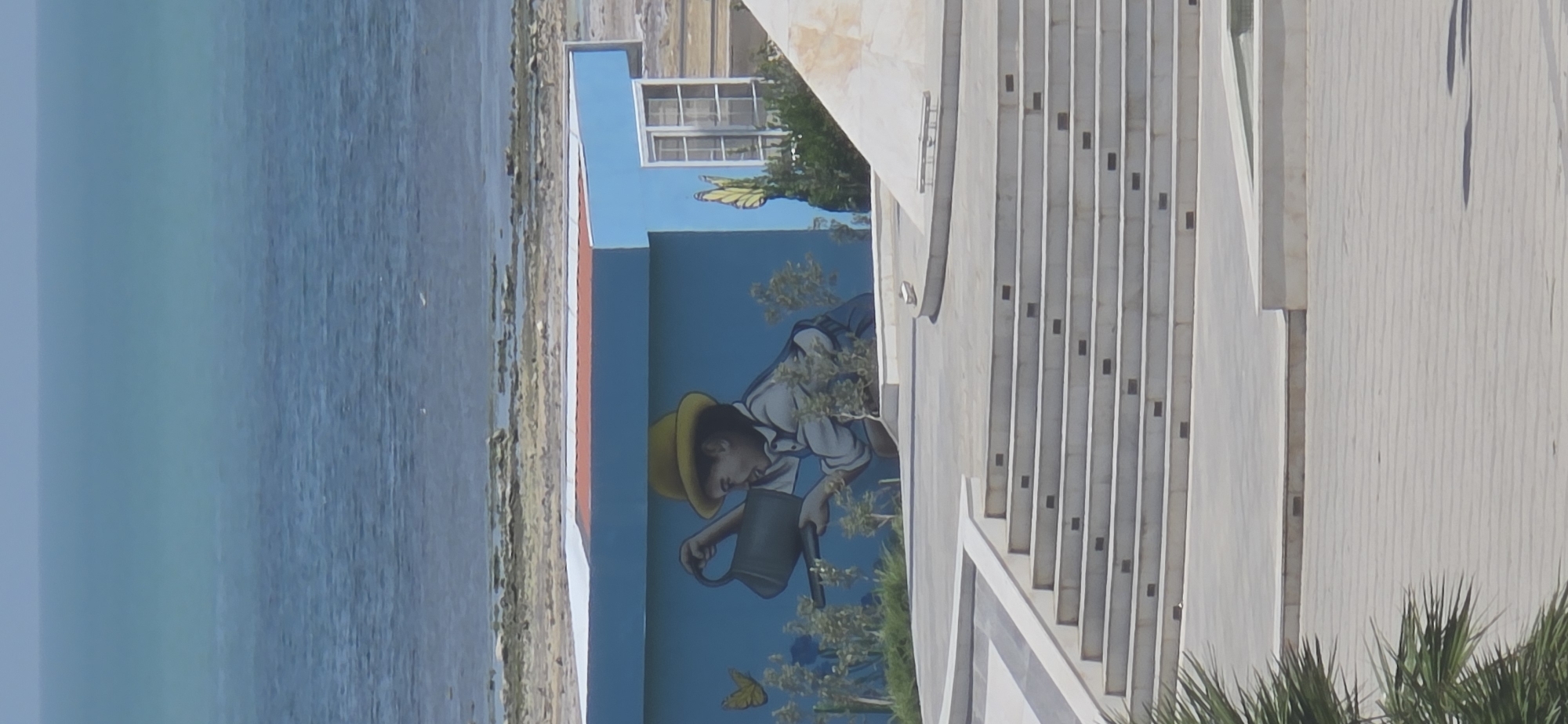 The image shows a mural painted on the side of a bright blue building. The mural depicts a young person wearing a yellow hat and a white shirt, holding a watering can and seemingly watering plants. Two yellow butterflies are also part of the mural. Behind the building, there is a body of water that appears to be the sea, with a rocky shoreline. In the foreground, there are concrete stairs and a paved area with some greenery. The overall scene suggests an outdoor recreational or public space, possibly near the coast. While the specific location in Sumgayit, Azerbaijan is not visually evident, the presence of a mural and public amenities could indicate such a setting.