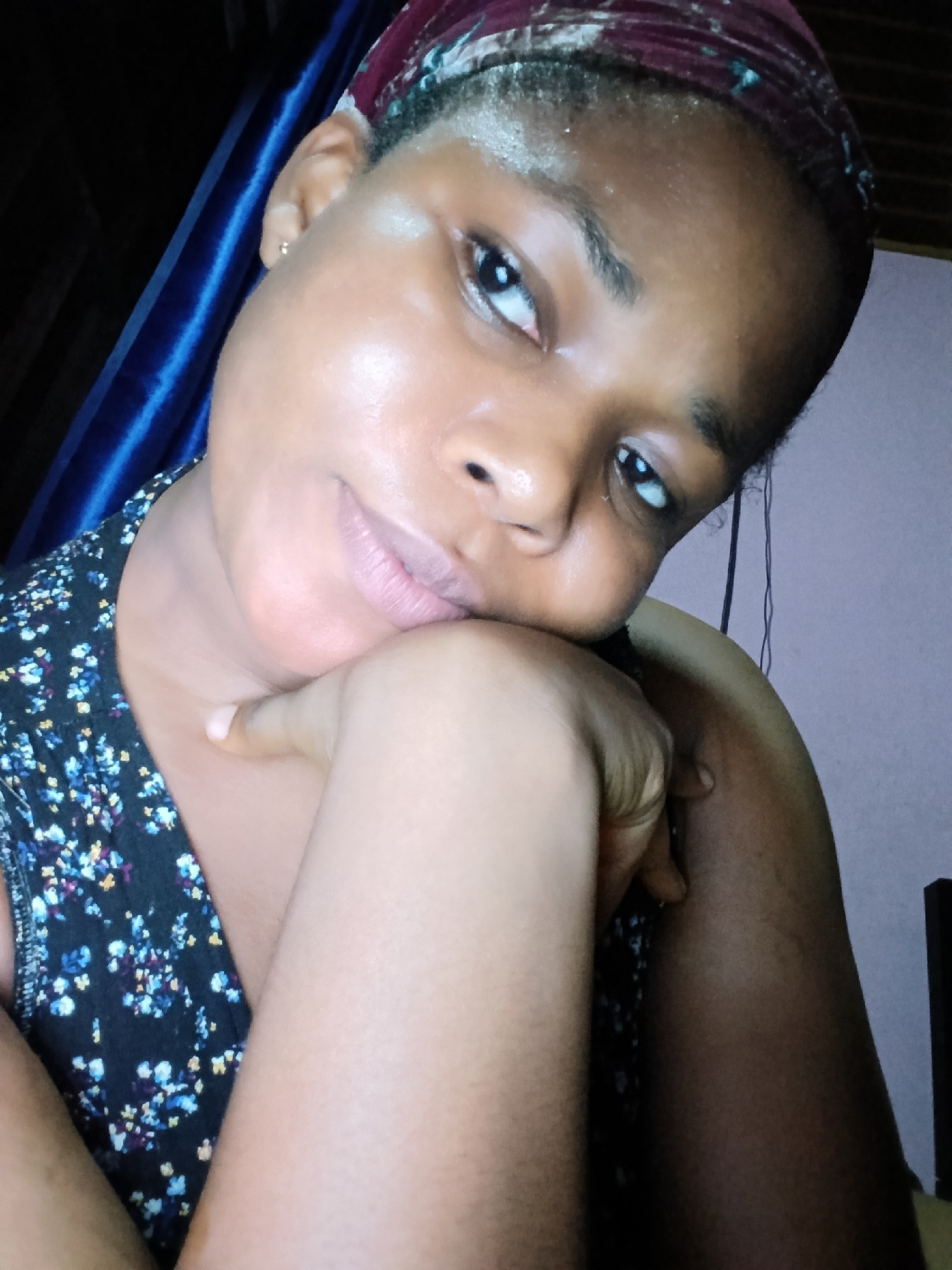 A young Black woman is posing for a selfie in Yenegoa, Nigeria. She is wearing a dark floral print top and a burgundy headscarf. Her left arm is bent, with her hand resting on her chin. The background shows a blue curtain and a plain wall. The lighting is dim, with a visible reflection on her forehead. The woman's expression is relaxed, with her eyes looking directly at the camera.