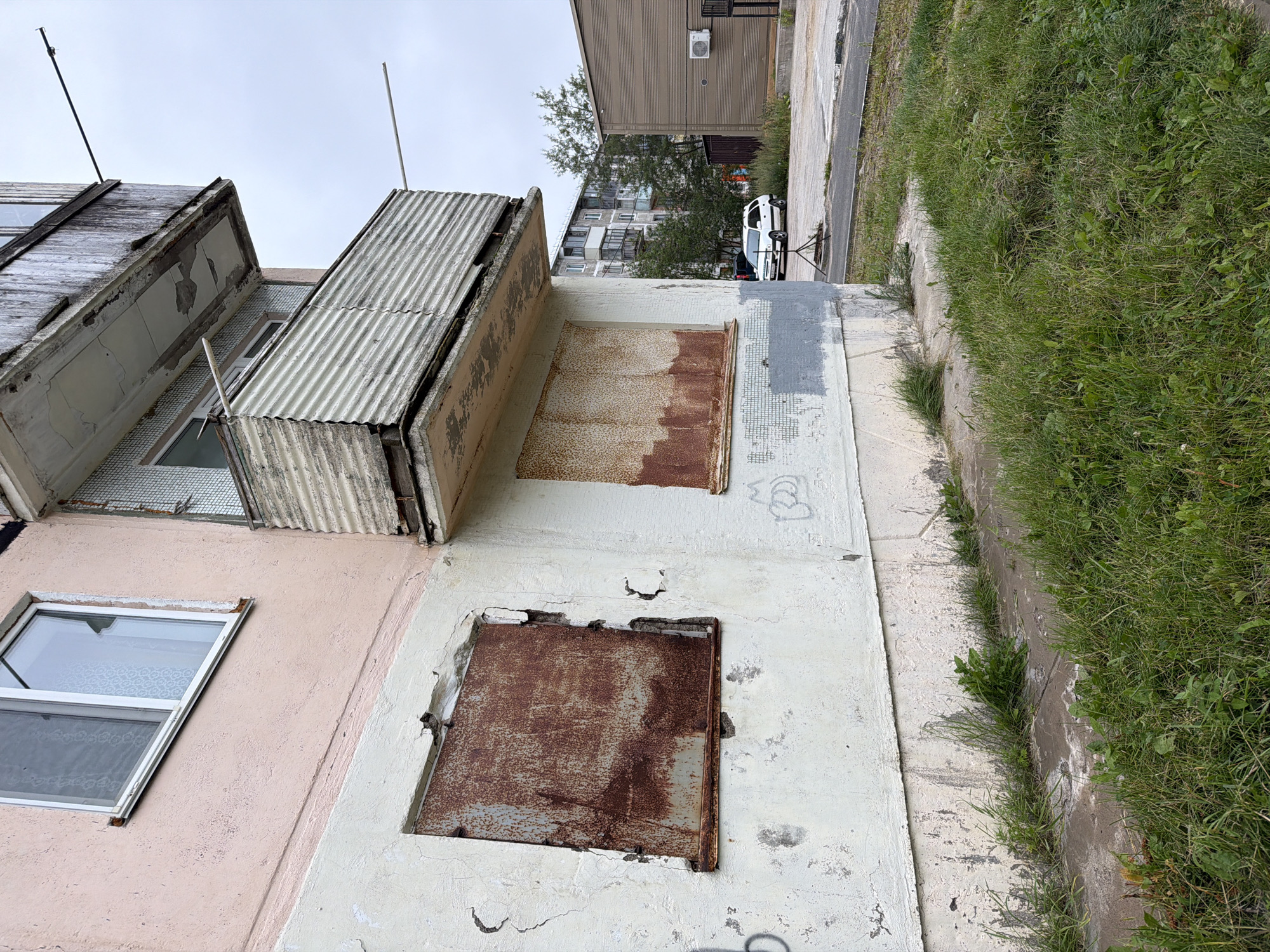 In Okha, Russia, an overcast sky looms over a weathered, multi-story building with a pink and white facade. The building displays signs of disrepair, with peeling paint and rusted metal coverings over some of its windows, particularly on the lower floors. A balcony on an upper level also shows wear and tear. In front of the building, overgrown grass and weeds line a cracked concrete walkway. Beyond the immediate building, a white car is parked near a modern, light brown building with an air conditioning unit mounted on its exterior. A taller apartment building and trees are visible in the background, suggesting a residential area.