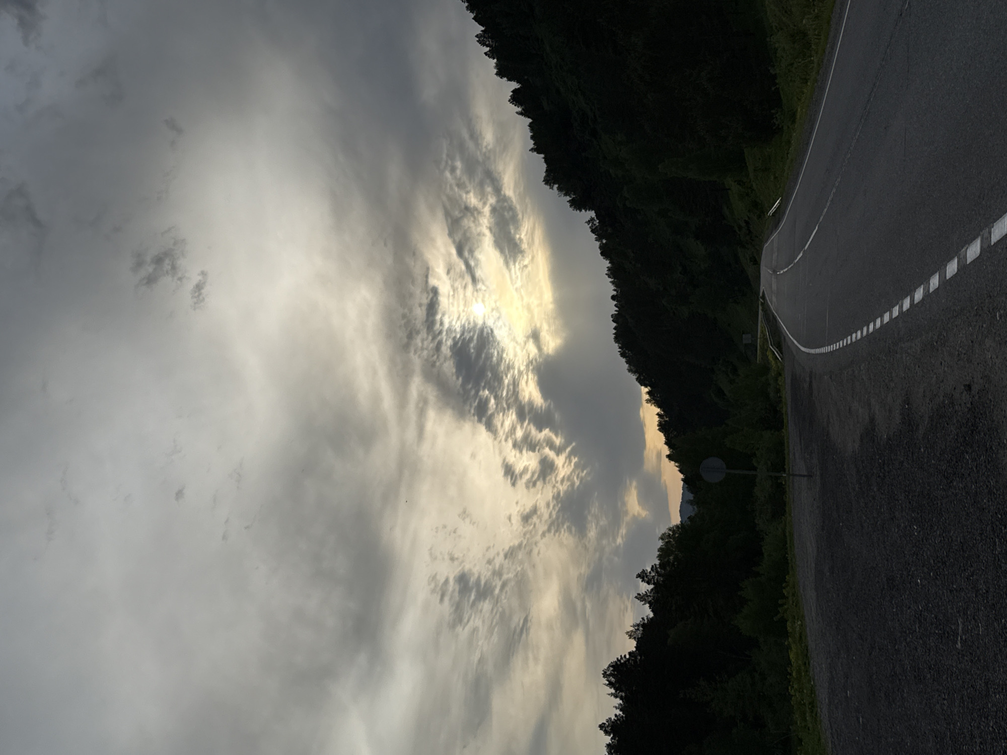 The image presents an upward-angled view of a road curving through a forest against a backdrop of a cloudy sky with the sun partially obscured. The road surface is dark with painted white lane markings. Dense trees border the road on both sides. The sky is filled with textured clouds, and the sun shines through a thin patch, creating a bright spot. The location is unknown and cannot be determined from the image.