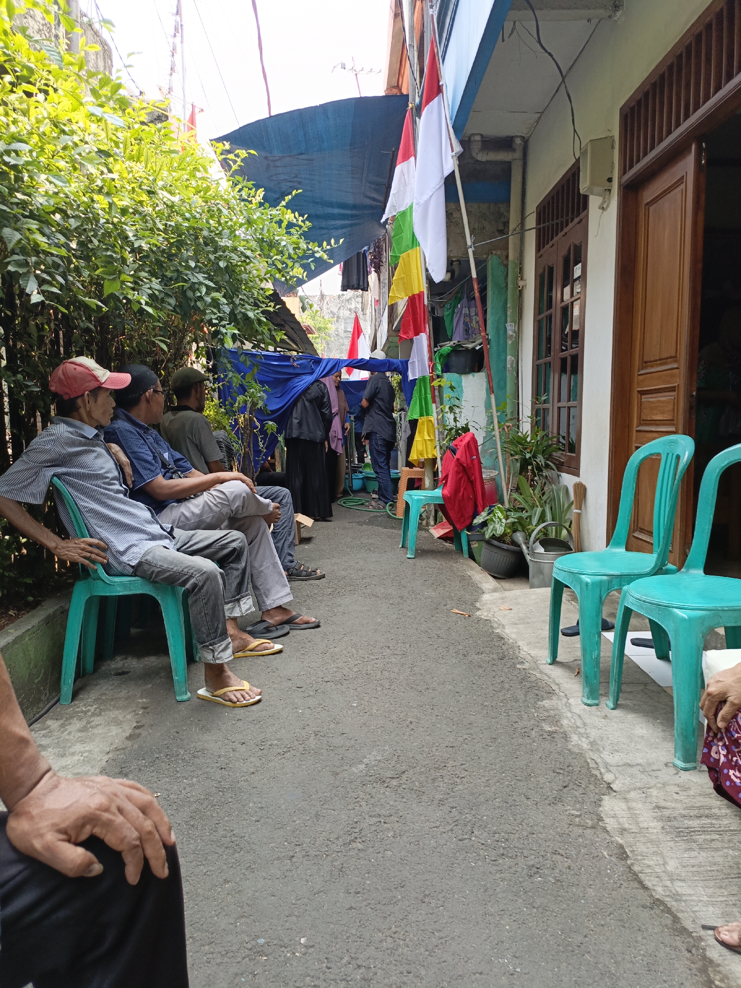 The image depicts an outdoor scene on a street, possibly in East Jakarta, Indonesia, as indicated by the presence of Indonesian flags. The setting appears to be a narrow alleyway or street in front of a residential building. Several individuals are seated on plastic chairs, facing away from the camera. The people are dressed casually. One man in the foreground wears a striped shirt, a red cap, and jeans. Next to him, another man is wearing a blue shirt and light-colored pants. Further down the row, two more men are seated, also facing away.
The environment includes dense green foliage on the left side of the image, which obscures part of the background. A blue tarp is visible overhead, providing shade. Indonesian flags, red and white, along with other colorful flags, are displayed on a pole in the middle ground. A building with wooden doors and windows is situated on the right side of the scene. There are several teal plastic chairs arranged on the pavement. Some plants in pots and a watering can are also present near the building. The ground is paved with asphalt and concrete. The overall impression is a community gathering or event taking place in a residential area.