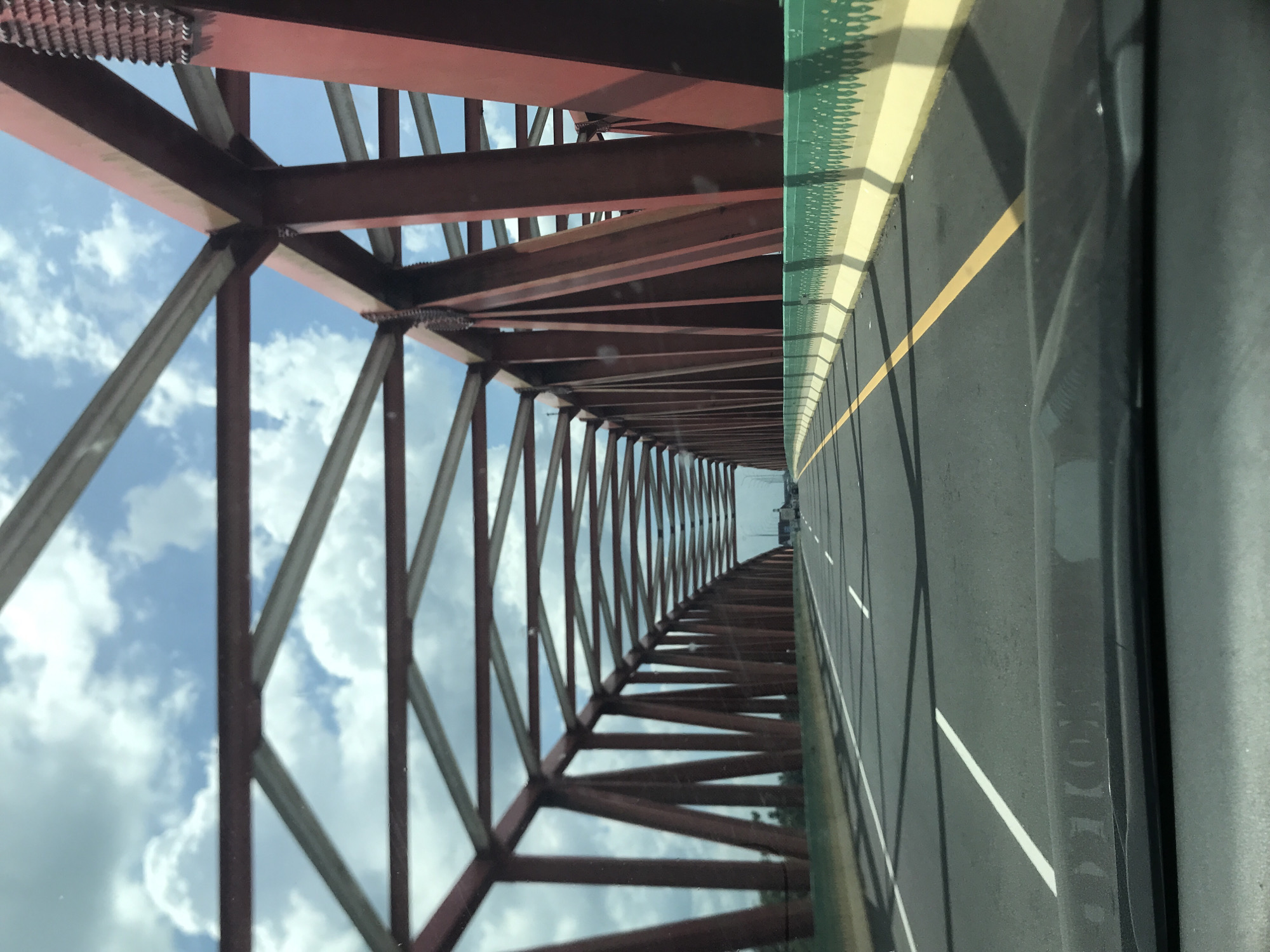 The image depicts a view from a vehicle moving along a bridge. The bridge's structure is made of red metal beams arranged in a geometric pattern against a cloudy sky. The road surface has white lane markings and a yellow dividing line. Part of the vehicle's interior is visible on the right side of the frame. The location is implied to be Gohor Lama, Indonesia, based on the prompt.