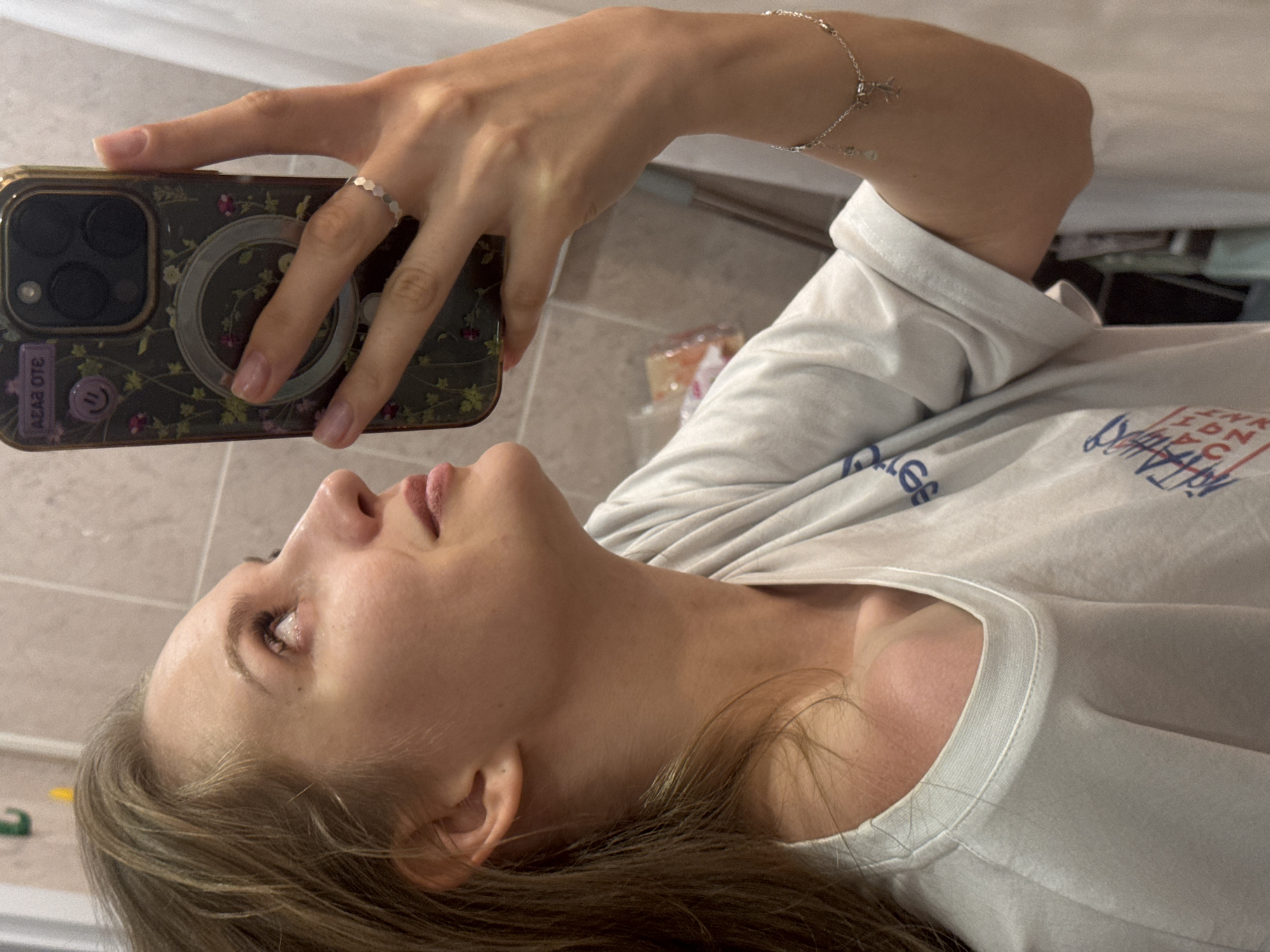 The image is a selfie. A fair-skinned woman with light brown hair is holding a smartphone with a patterned case, taking the picture from a low angle. She is wearing a light-colored t-shirt. A silver ring is visible on her finger, and a silver bracelet on her wrist. The background suggests a bathroom setting, with light-colored tiled walls. The location in Izhevsk, Russia is not discernible from the image.