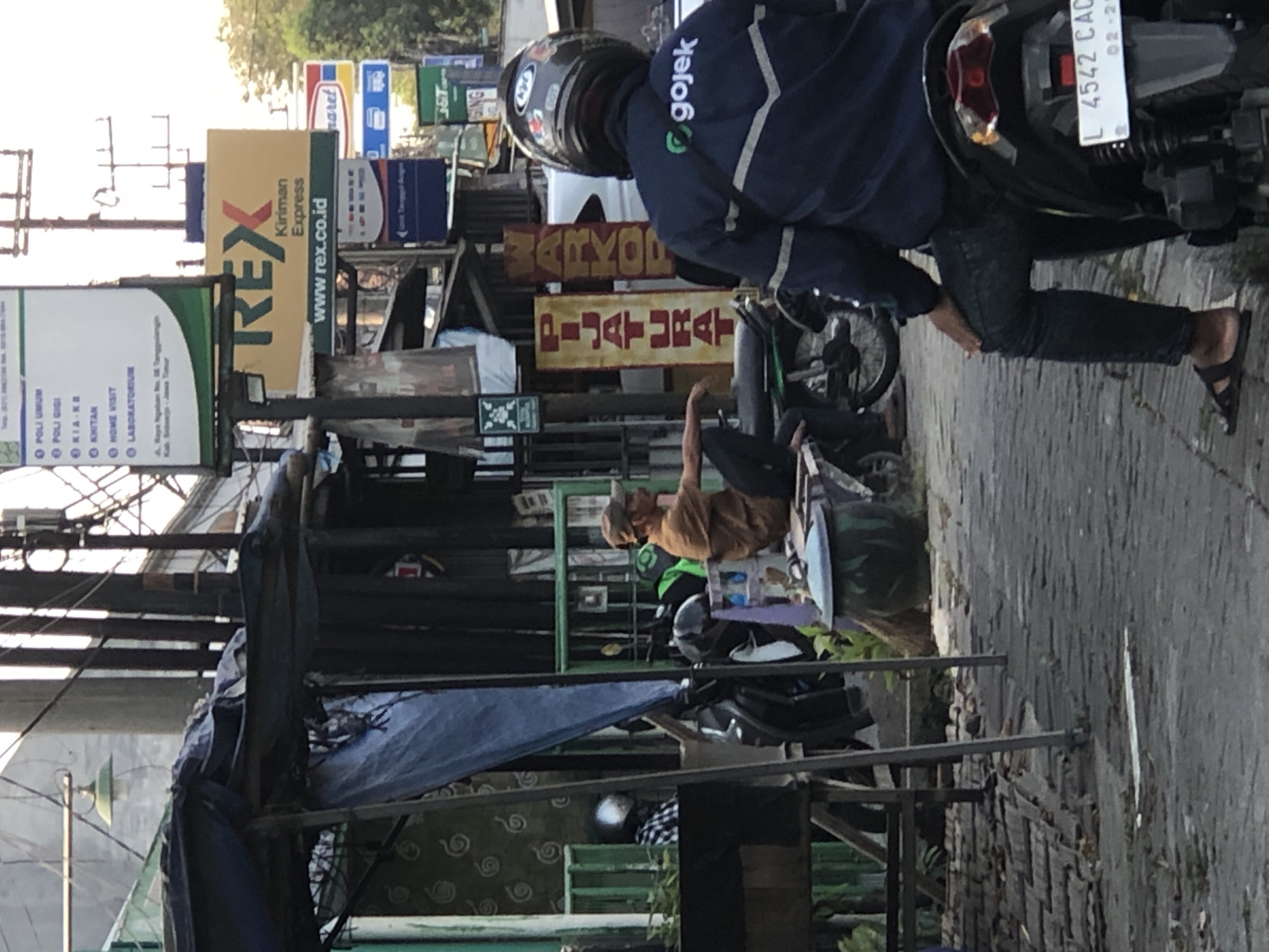 The image captures a street scene in Ngaban, Indonesia, with a focus on a man seated on a motorcycle, wearing a navy blue jacket with the "gojek" logo. He is facing away from the camera, and his legs are crossed. To his left, another man wearing a tan shirt and a cap is seated on a bench, appearing to be resting or waiting. Several motorcycles are parked in the background, and a blue tarp covers a stall. Signs for "REX Kiriman Express" and other businesses are visible on buildings in the background. The ground is paved with stones, and there are various objects and structures, including a trash can, a broom, and a street lamp, indicative of an urban environment. The lighting suggests it is daytime.