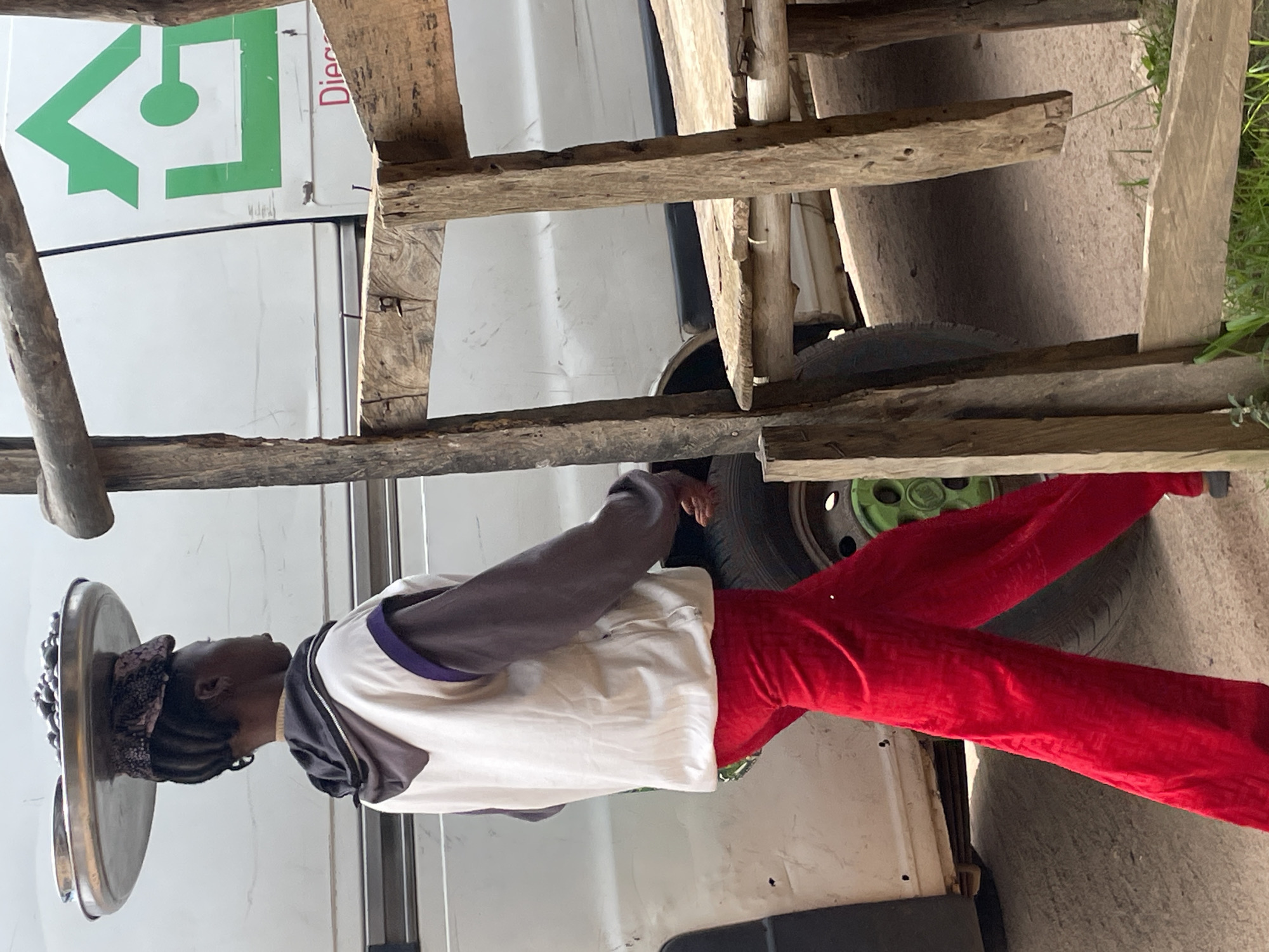 A person is walking on a dirt road in the city of Wang, Nigeria, carrying a metal tray of food on their head. The person is wearing a white vest with purple trim over a dark hoodie, and red trousers. Behind the person is a white van with a green house logo on the side. To the right of the person is a rustic wooden structure with a chair. The ground is dirt and small stones. The lighting suggests it is daytime.