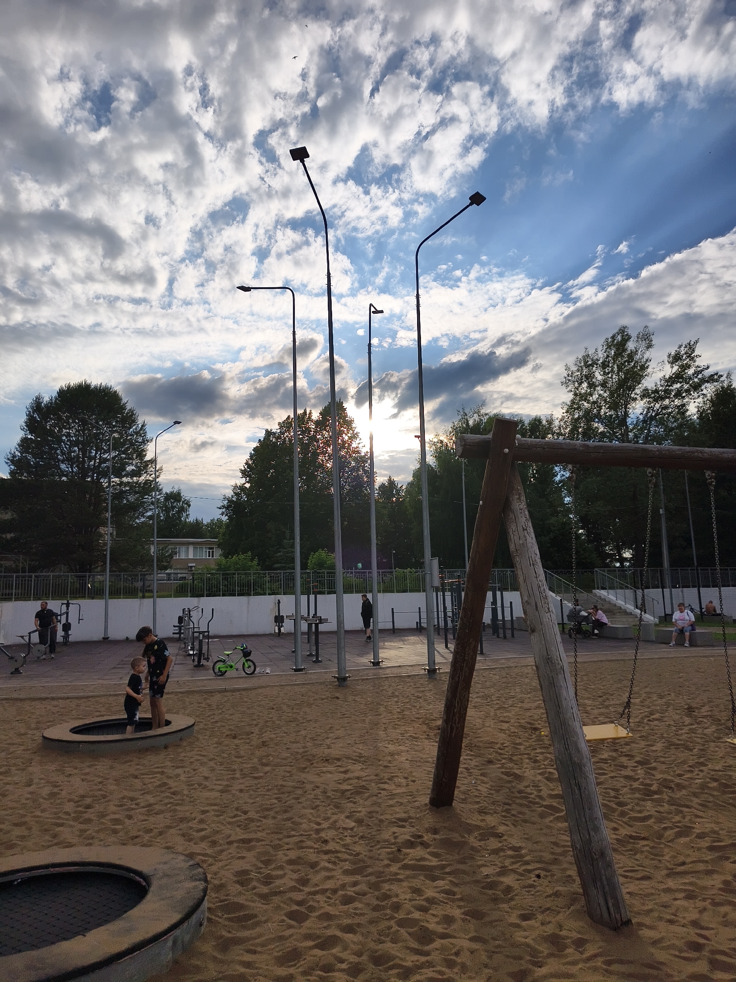The image depicts an outdoor recreational area in Chaikovsky, Russia, featuring a playground with a sandpit, swings, and exercise equipment. Two children are standing in a sunken trampoline in the foreground. In the background, several adults are seen using the exercise machines and sitting on benches. The sky is partially cloudy with the sun peeking through, casting a warm glow. Trees and buildings are visible in the distance, indicating an urban or suburban setting. The overall scene suggests a community space for recreation and exercise.