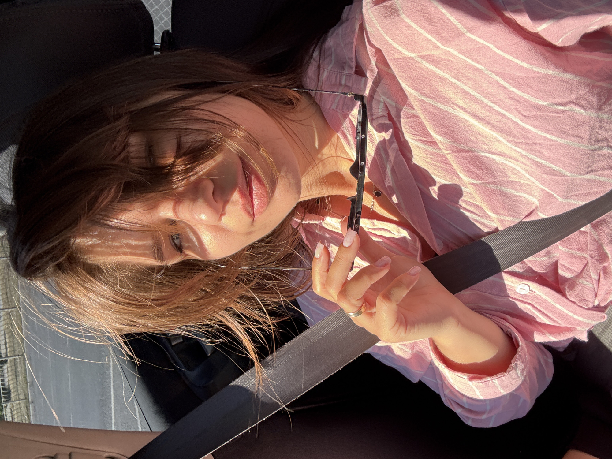 This is a close-up, rotated media file of a woman in a car, likely a selfie taken by herself. She has long, brown hair that is falling across her face, partially obscuring her features. She is wearing a pink and white striped button-up shirt and appears to be holding a pair of glasses in her right hand. Her left hand is visible, with a ring on her finger. The sunlight is casting strong shadows across her face and the shirt, indicating it is daytime. The background suggests she is in the passenger seat of a car, with a seatbelt clearly visible across her chest. The scene appears relaxed and casual, with the woman's expression suggesting a peaceful or thoughtful mood. There is no visible text in the media file.