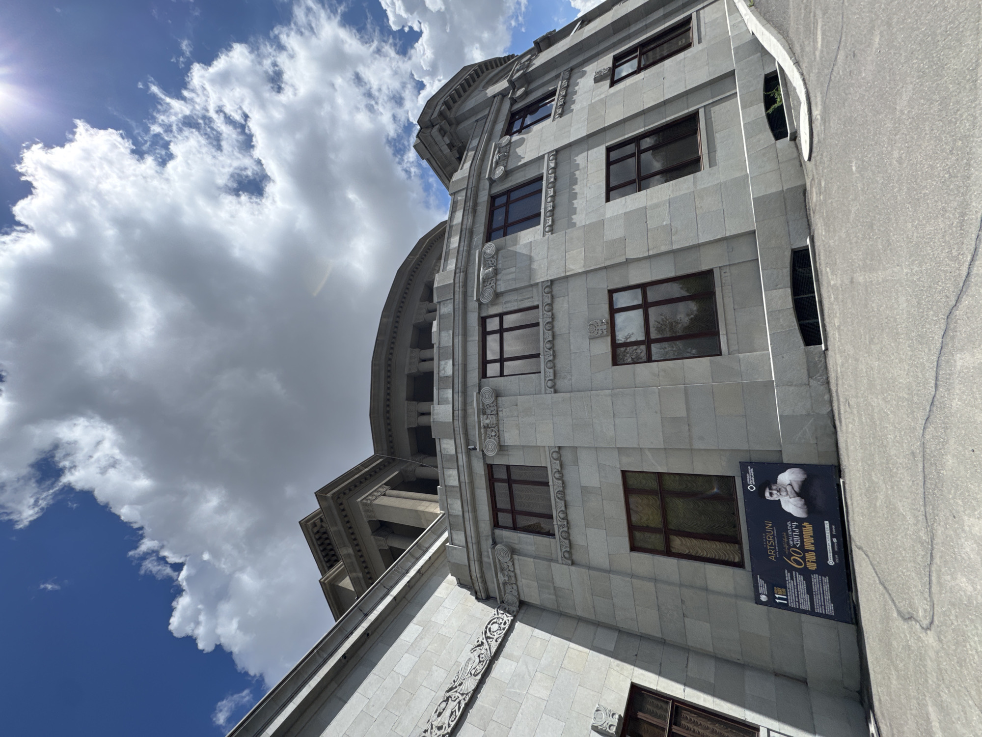 This image captures a grand, light-colored stone building in Yerevan, Armenia, seen from a low-angle perspective. The building exhibits ornate architectural details, including intricate carvings, decorative columns, and windows with dark frames, suggesting a classical or Art Deco style. Its facade is curved, adding to its distinctive appearance. The sky above is a vibrant blue with large, white, fluffy clouds, and a sun flare is visible in the upper left corner, indicating a bright, sunny day.
Attached to the building's exterior, near the paved ground, is a black promotional banner. The banner features a black and white image of a man, likely related to the event advertised. Visible text on the banner includes "ARTSRUNI" in large English letters, followed by "CONCERT" and "STATE CHOIR OF ARMENIA." It also displays the number "60" and Armenian text that translates to "anniversary," "Komitas," "named after," "national," "musical," and "center," suggesting a 60th-anniversary concert by the State Choir of Armenia, possibly at the Komitas National Musical Center. The date "11 ՄԱՅԻՍ" (May 11) and time "19:00" are also present, along with a QR code and social media icons.
The scene is static, devoid of human activity, focusing instead on the impressive architecture and the clear, bright weather. The overall impression is one of a significant cultural or governmental building under a beautiful sky.