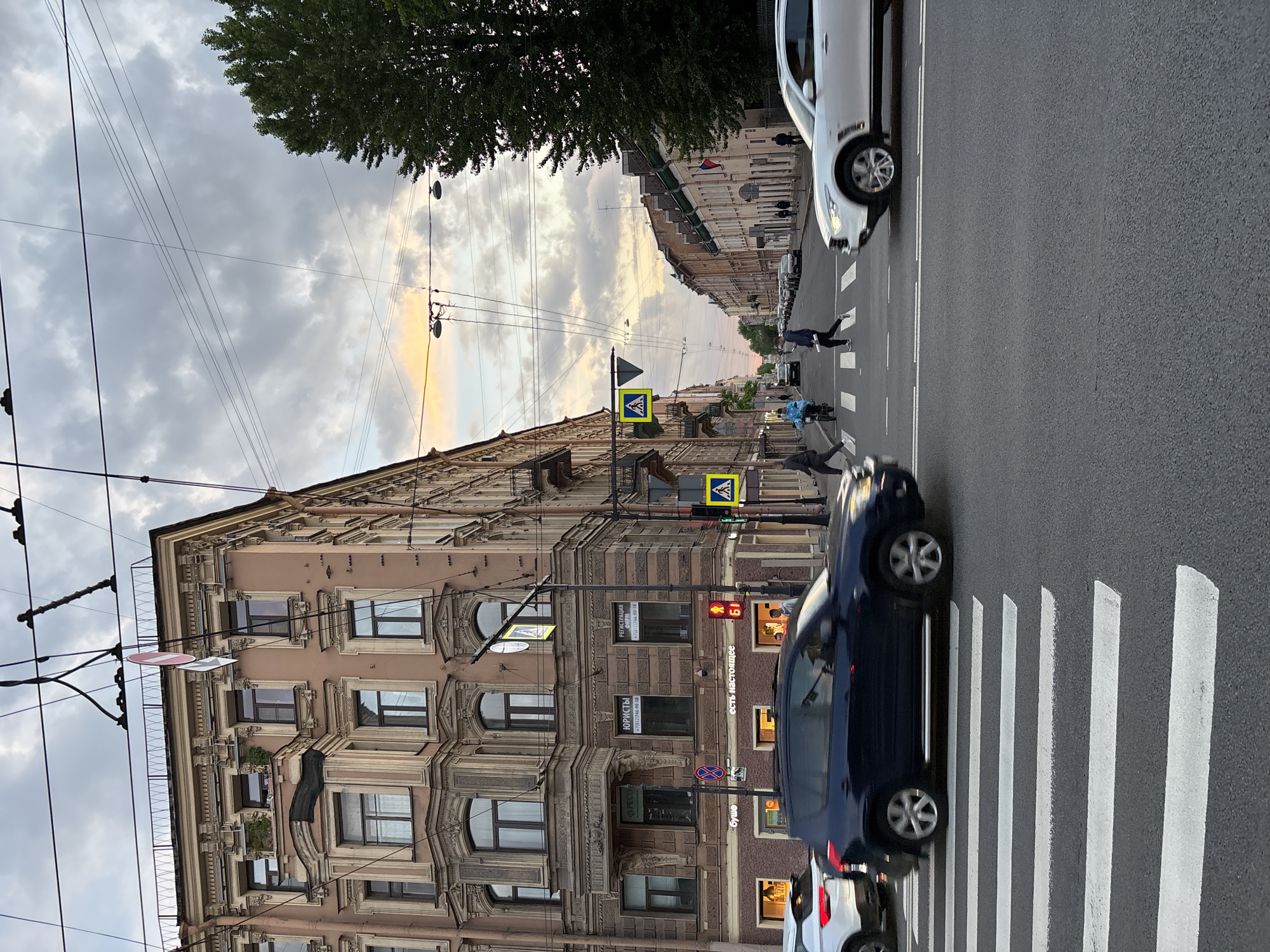 The image depicts a bustling street scene in Saint Petersburg, Russia, likely during the late afternoon or early evening given the soft, warm light in the cloudy sky. The street is characterized by a prominent crosswalk in the foreground and is flanked by tall, ornate classical-style buildings on one side. A network of overhead wires, possibly for trams or trolleybuses, spans the street.
Several subjects populate the scene:
- **Vehicles:** A dark blue car is positioned on the crosswalk, with a white car visible behind it. Another white car is further down the street on the right.
- **People:** Multiple pedestrians are using the crosswalk. One person in dark attire is walking across, while another in a blue jacket appears to be on a scooter or bicycle. More pedestrians can be seen further along the street.
- **Objects:** Street signs include yellow diamond-shaped pedestrian crossing indicators and a red circular "No Stopping" sign. A red traffic light shows a numerical countdown of "61." A tree with green foliage is visible against the sky.
The activity revolves around typical urban transit, with vehicles and pedestrians navigating the street. The buildings showcase detailed architecture with multiple windows and decorative elements.
Visible Russian text on the building facades includes "ЮРИСТЫ" (Lawyers) and "РЕГИСТРАЦИЯ ФИРМ" (Company Registration), accompanied by a local Saint Petersburg phone number (+7(812)746-90-18). Other text reads "буше" (Bushe, possibly a chain like a bakery/cafe) and "есть настоящее" (meaning "there is real" or "there is the present," potentially a slogan). The text "PECO" is also partially visible.