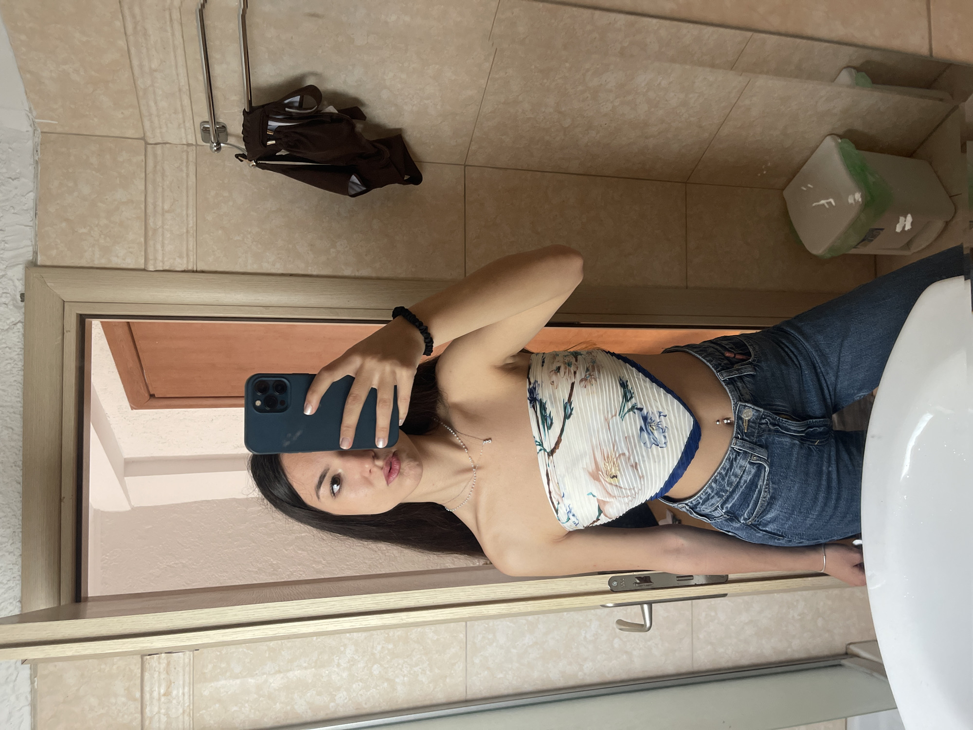 A woman is taking a selfie in a bathroom. She is wearing a floral tube top and jeans. The bathroom has beige tile walls and a wooden door frame. There is a sink and a toilet in the bathroom. A bag is hanging on the wall. The location is likely in Nea Makri Municipal Unit, Greece, although there is no verifiable information to confirm this.