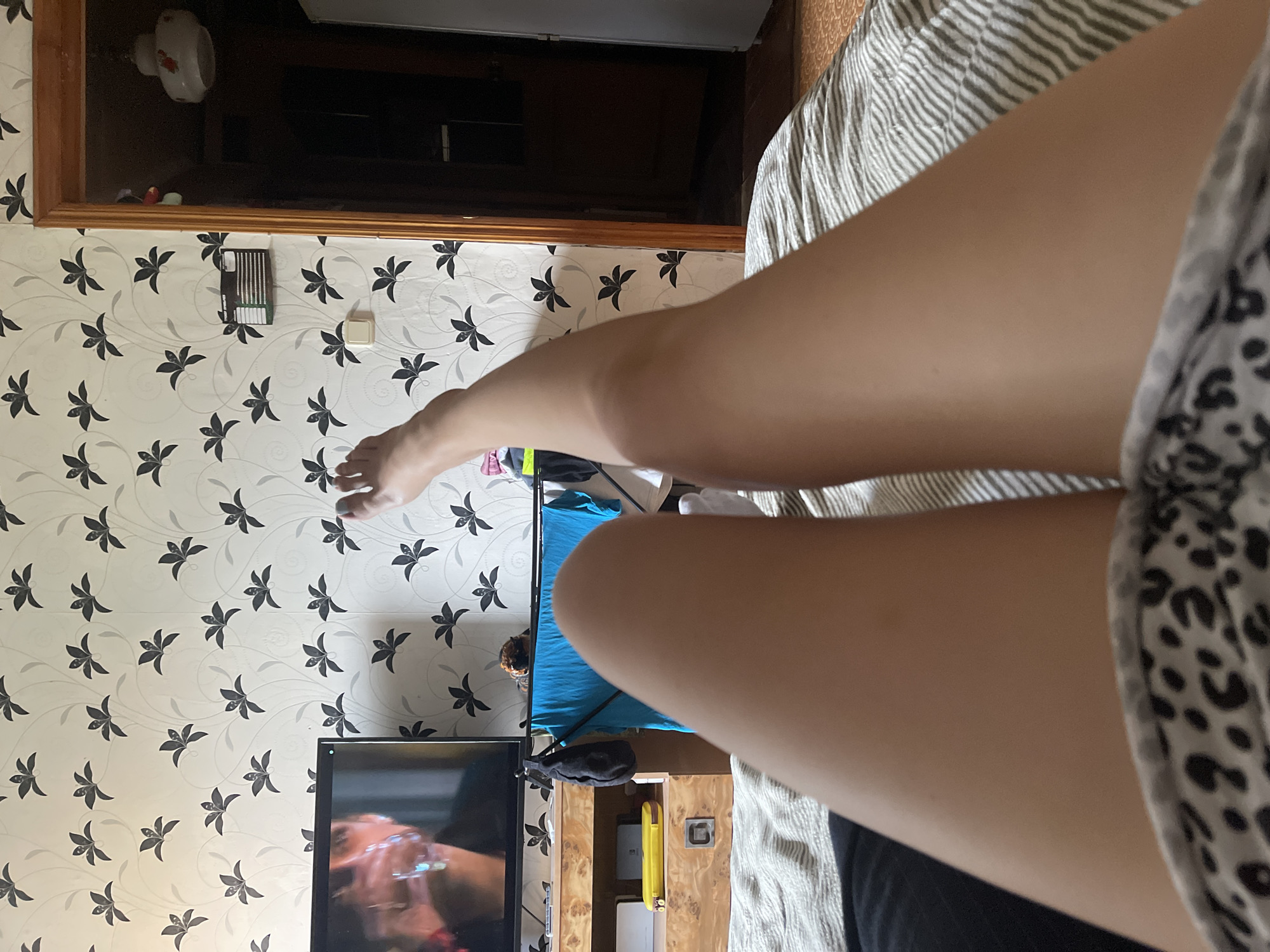 The image depicts a person's legs and feet from a first-person perspective, lying on a bed. The room has floral wallpaper and a television is visible on a wooden stand. There is also a doorway leading to another room, a clothes drying rack with blue clothing, and a refrigerator. The scene is located in Shcherbinka, Russia.