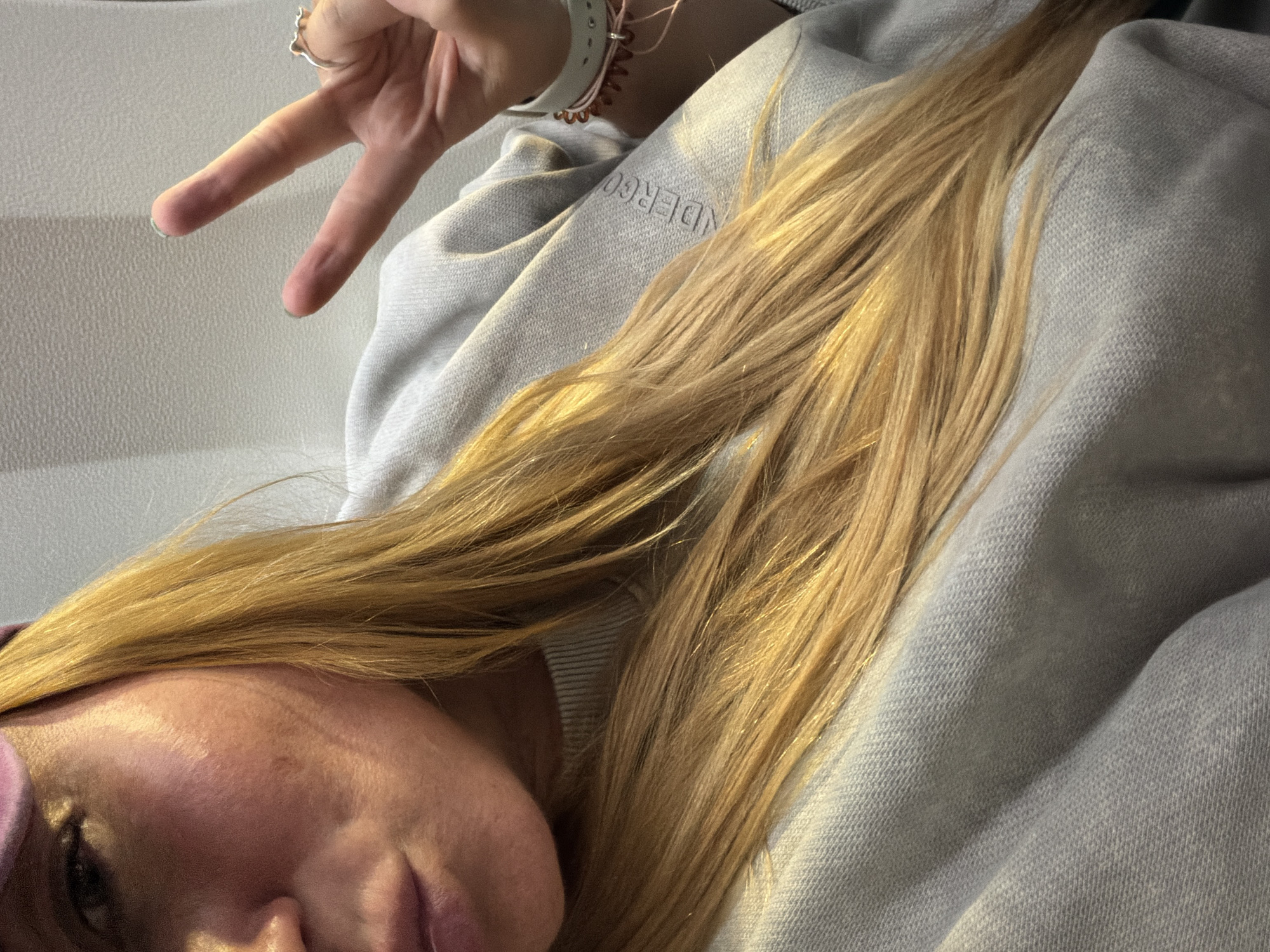A selfie-style image shows a person with long, light blonde hair lying against a gray, textured surface, possibly a garment. The person's face is partially visible, showing part of their forehead, nose, and lips. They are making a peace sign with their hand. A ring is visible on their finger. The light is warm and illuminates their hair. The setting is indeterminate. There is no evidence to suggest the location is in Madrid, Spain.