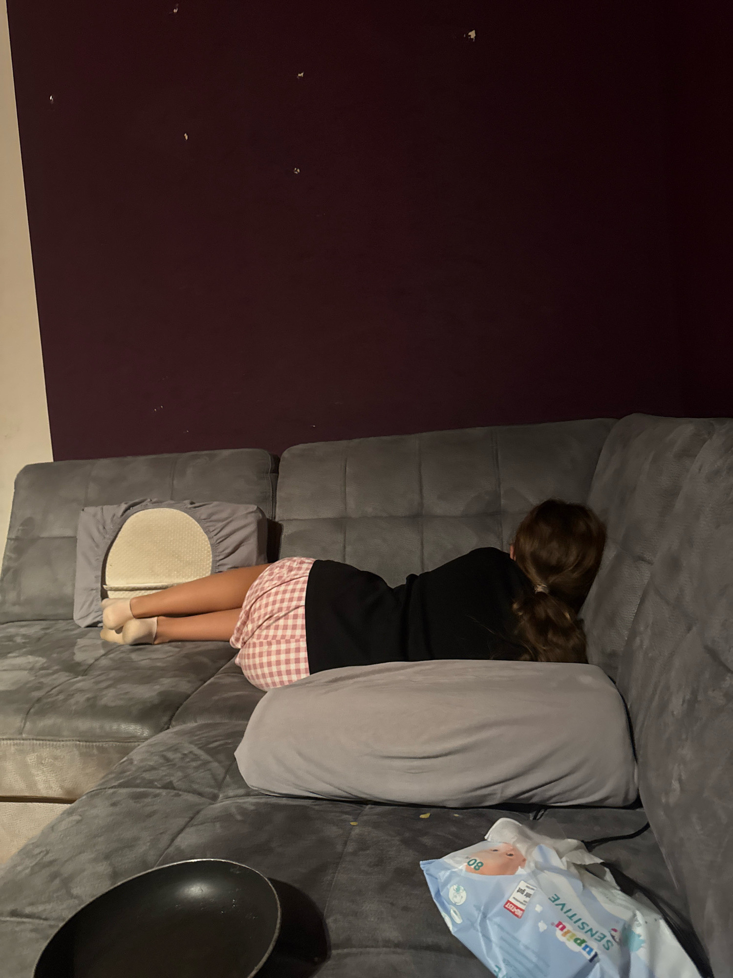 A young person is lying on a grey sectional sofa, facing away from the camera, with their head resting on a grey pillow. The person is wearing a black long-sleeved shirt and pink and white checkered shorts. Their legs are bent, and they are wearing light-colored socks. A cream-colored pillow with a textured circular pattern is visible behind their legs. To the right of the person, a grey pillow is placed horizontally on the sofa. In the foreground, a black frying pan is visible on the left side of the sofa. To the right of the frying pan, a package of baby wipes is on the sofa. The wall behind the sofa is a dark purple color with several small light-colored specks. The overall scene appears to be indoors, likely in a residential setting. There is no visual information in the image to suggest a specific location within the city of Überherrn, Germany.