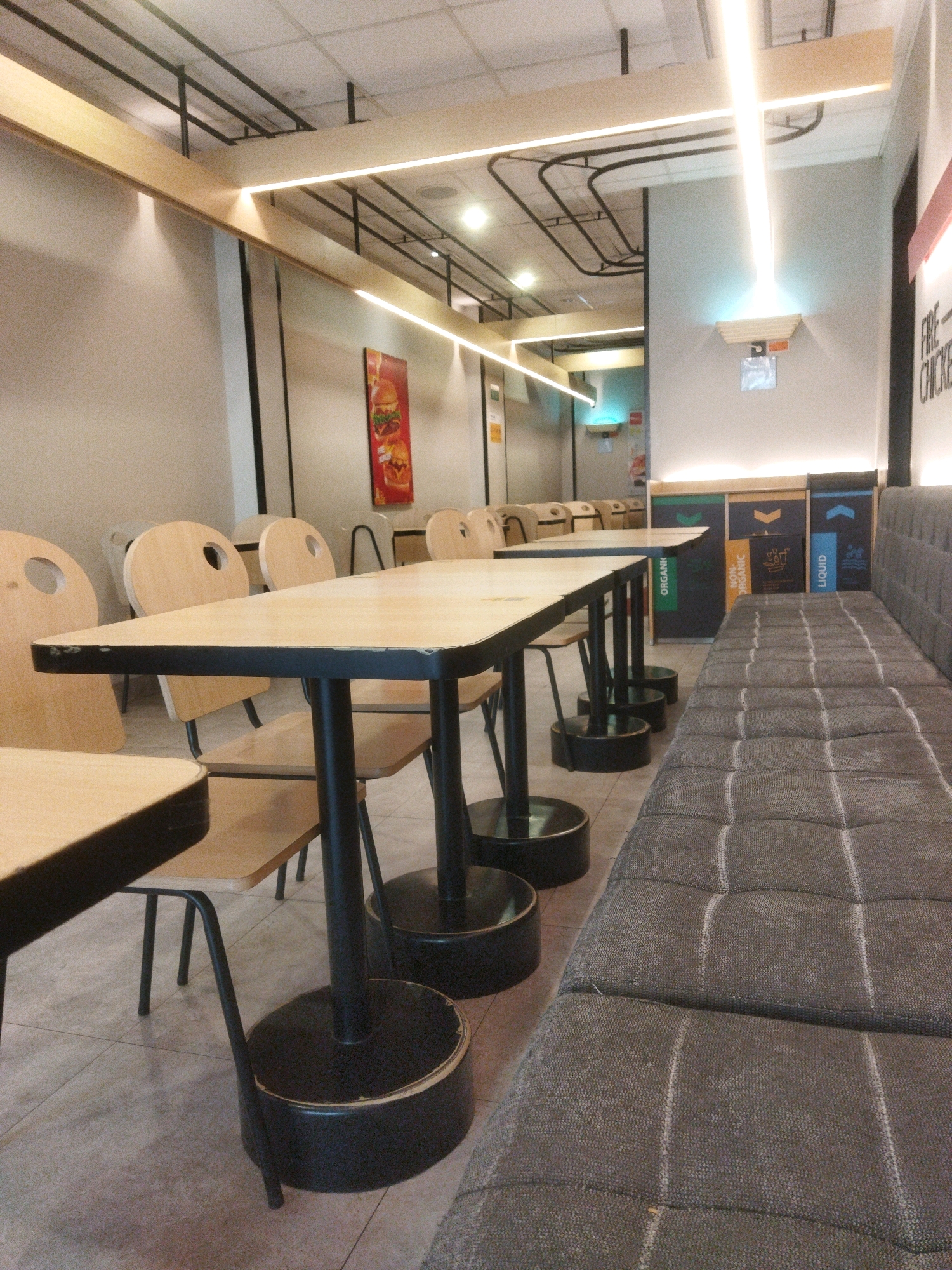 The image depicts the interior of a fast-food restaurant with a modern, minimalist design. The dining area features light-colored wooden tables with black metal bases and matching chairs, some of which have circular cutouts in the backrests. A long, gray upholstered bench with a tufted pattern runs along the right side of the room. In the background, there are three recycling bins with labels indicating "ORGANIC," "NON-ORGANIC," and "LIQUID." The walls are painted a light neutral color, and the ceiling has exposed industrial elements and linear lighting fixtures. A framed poster of a burger is visible on the wall. The overall ambiance suggests a clean and contemporary dining space. The image does not provide any specific information about the city of Pasuruan, Indonesia, or any individuals present in the scene.