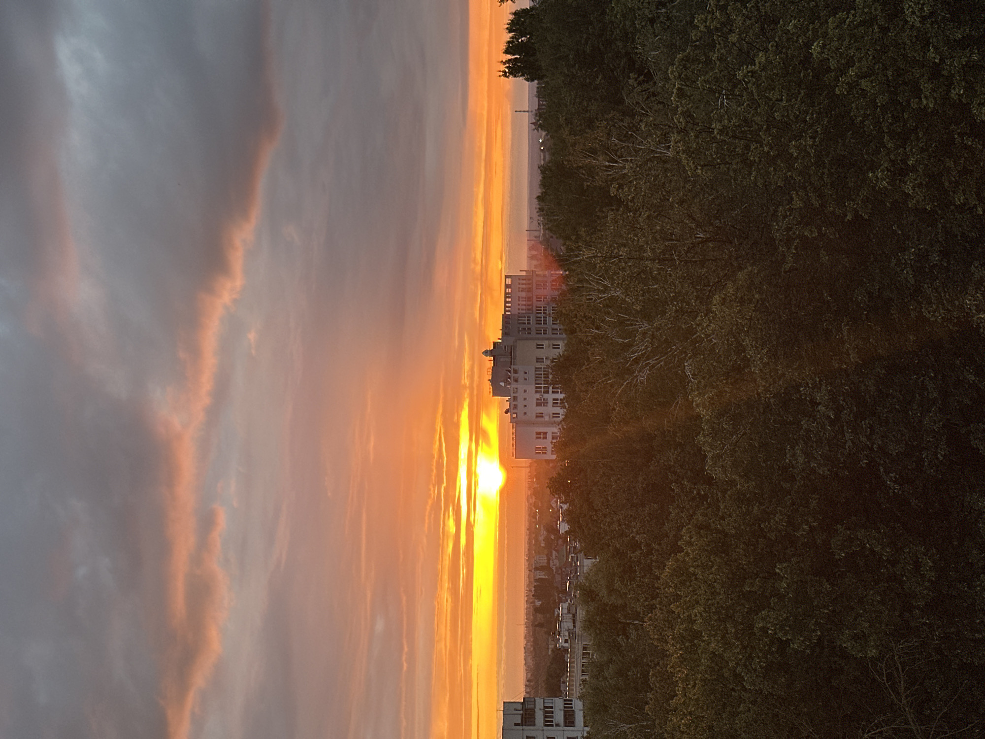 The image depicts a cityscape during sunset, viewed from an elevated position. The sky is dominated by a brilliant orange and yellow sunset, with streaks of pink and gray clouds stretching across. In the foreground, a dense canopy of dark green trees occupies the lower portion of the frame, obscuring much of the ground.
Emerging from behind the trees are several multi-story buildings. To the left, a lighter-colored building with multiple windows is visible. To the right and slightly further back, a larger, white building with a modern architectural design stands out. It features numerous windows and a flat roof. In the distance, a hazy horizon is dotted with what appear to be industrial structures or smokestacks, suggesting the presence of an industrial area or a larger city beyond. The overall impression is of a transitional zone between a natural environment and urban development, captured at the dramatic transition of day to night. The location is identified as Krugloye, Russia.