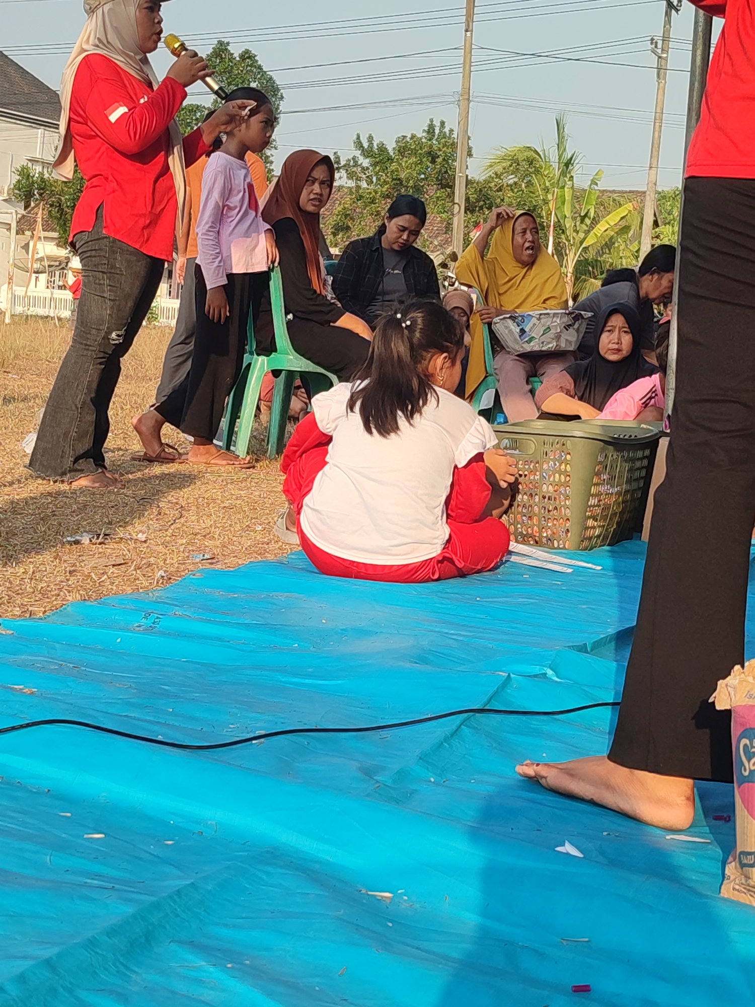 In Nusa Jaya, Indonesia, a group of people are gathered outdoors, possibly for an event or performance. In the foreground, a young girl in a white shirt and red pants sits on a blue tarp, facing away from the camera. Next to her, a green plastic chair holds a large green laundry basket filled with items. Behind the girl, a woman in a red shirt and hijab stands holding a microphone, appearing to speak. To her right, a young girl in a lavender shirt stands looking to the side. Other individuals, including women in hijabs, are seated or standing in the background, some engaged in conversation. The environment appears to be a dry, grassy area with trees and residential structures visible in the distance, under a bright sky.