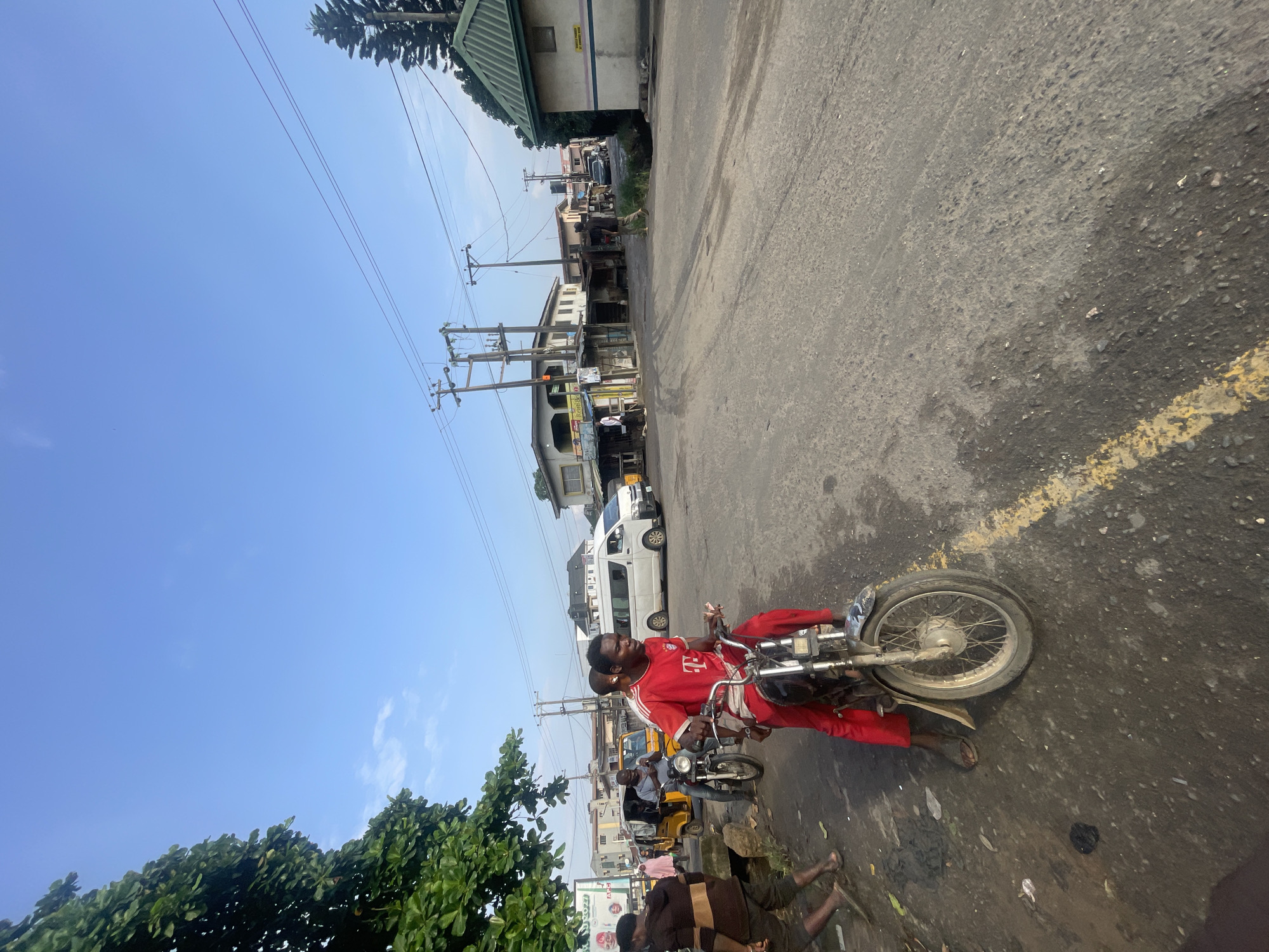 A man in a red shirt and pants rides a motorcycle in Akowonjo, Nigeria. The motorcycle has a chrome-colored exhaust pipe and silver spokes. The man's left leg is bent at the knee, and his right foot is resting on the ground. He is holding the handlebars with both hands, and his gaze is directed forward. The motorcycle's front tire is visible, and it is a spoked wheel with a rubber tire. The road is paved with asphalt and is worn and cracked. A yellow line is painted on the road. In the background, there are several buildings, including a white van, a yellow auto-rickshaw, and other vehicles. Trees are also visible in the background. The sky is blue with a few white clouds. The scene appears to be a street in a busy urban area.