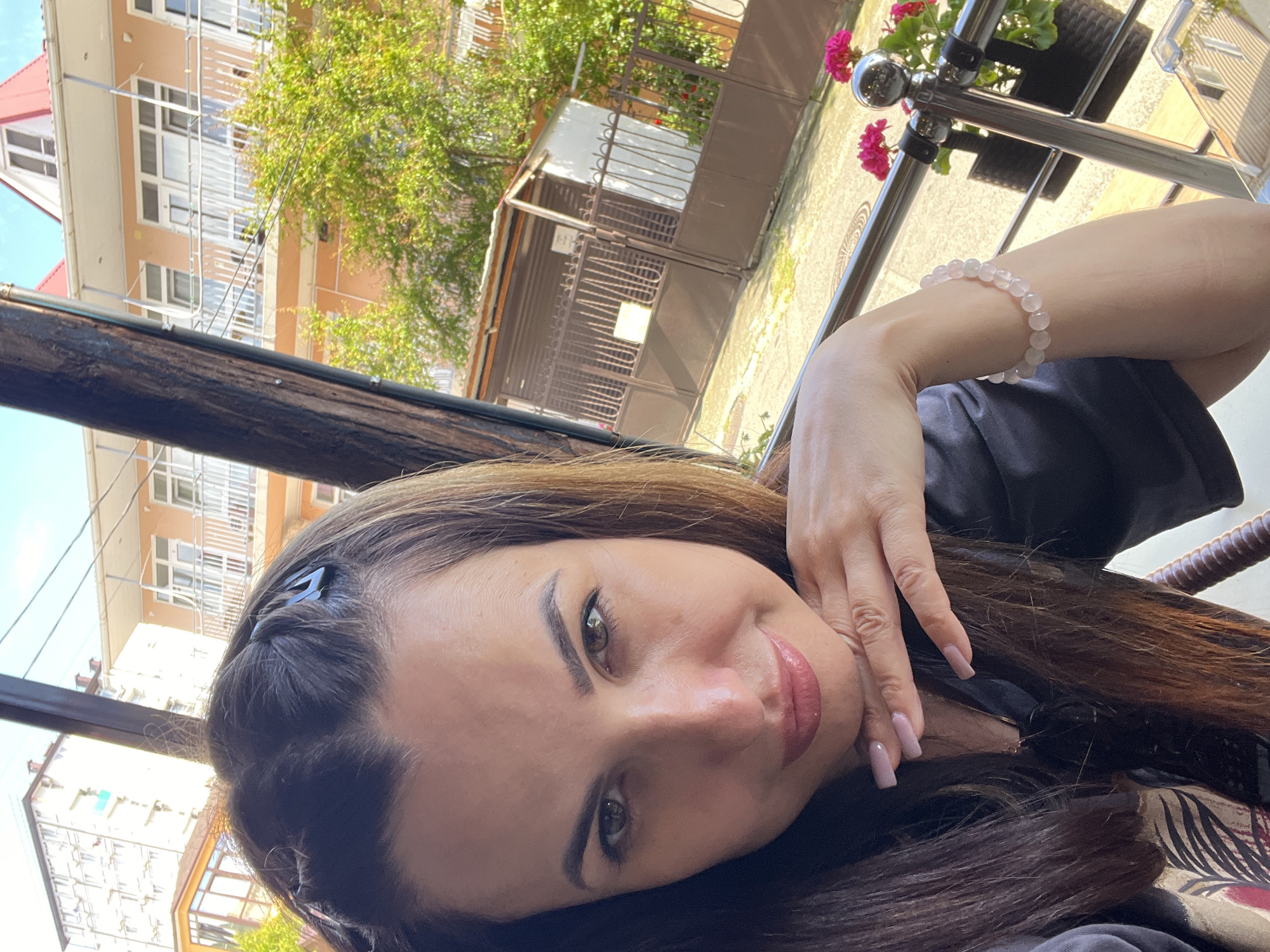 In a selfie taken outdoors, a woman with long brown hair, some of which is styled with two braids at the front, looks directly at the camera. She is resting her chin on her right hand. Her fingernails are painted a light pink, and she is wearing a dark top. On her left wrist, she wears a bracelet made of light pink beads. Behind her, a building with multiple floors and balconies is visible, along with green trees. A metal railing separates the foreground from the background, and on it, a potted plant with pink flowers is visible. The overall setting appears to be a balcony or terrace in a city.