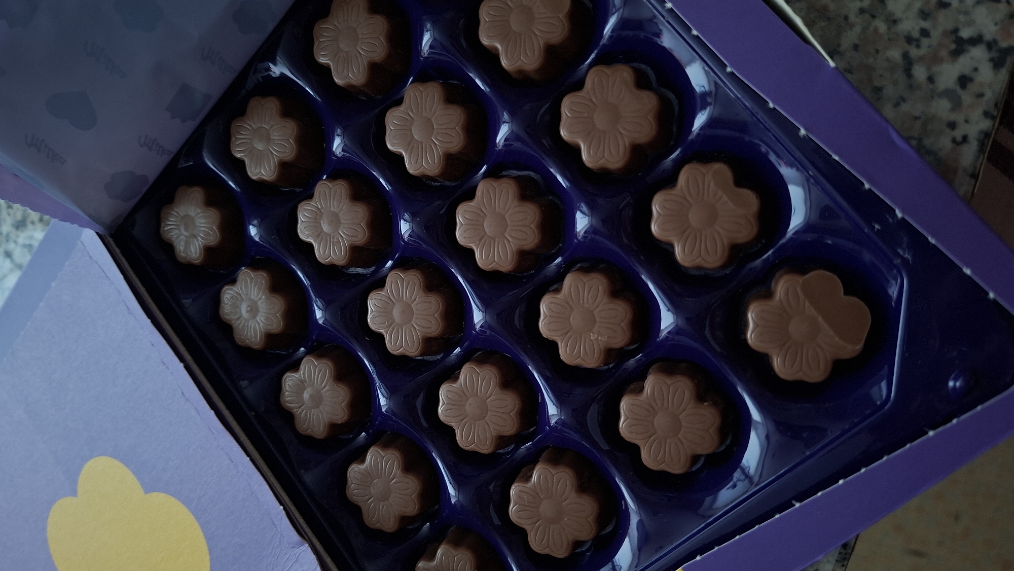 A box of chocolates is shown in an overhead shot. The box is purple and appears to be made of cardboard. Inside the box, the chocolates are arranged in a grid of six rows and four columns, with a total of 24 chocolates. Each chocolate is shaped like a flower and is made of milk chocolate. The chocolates are placed in individual purple plastic molds. The lid of the box is partially open and shows a purple paper with a yellow cloud-shaped cutout on the left and a pattern of clouds and hearts on the right. The background appears to be a countertop. The location in the city of Myadzyel, Belarus, is not determinable from the image.