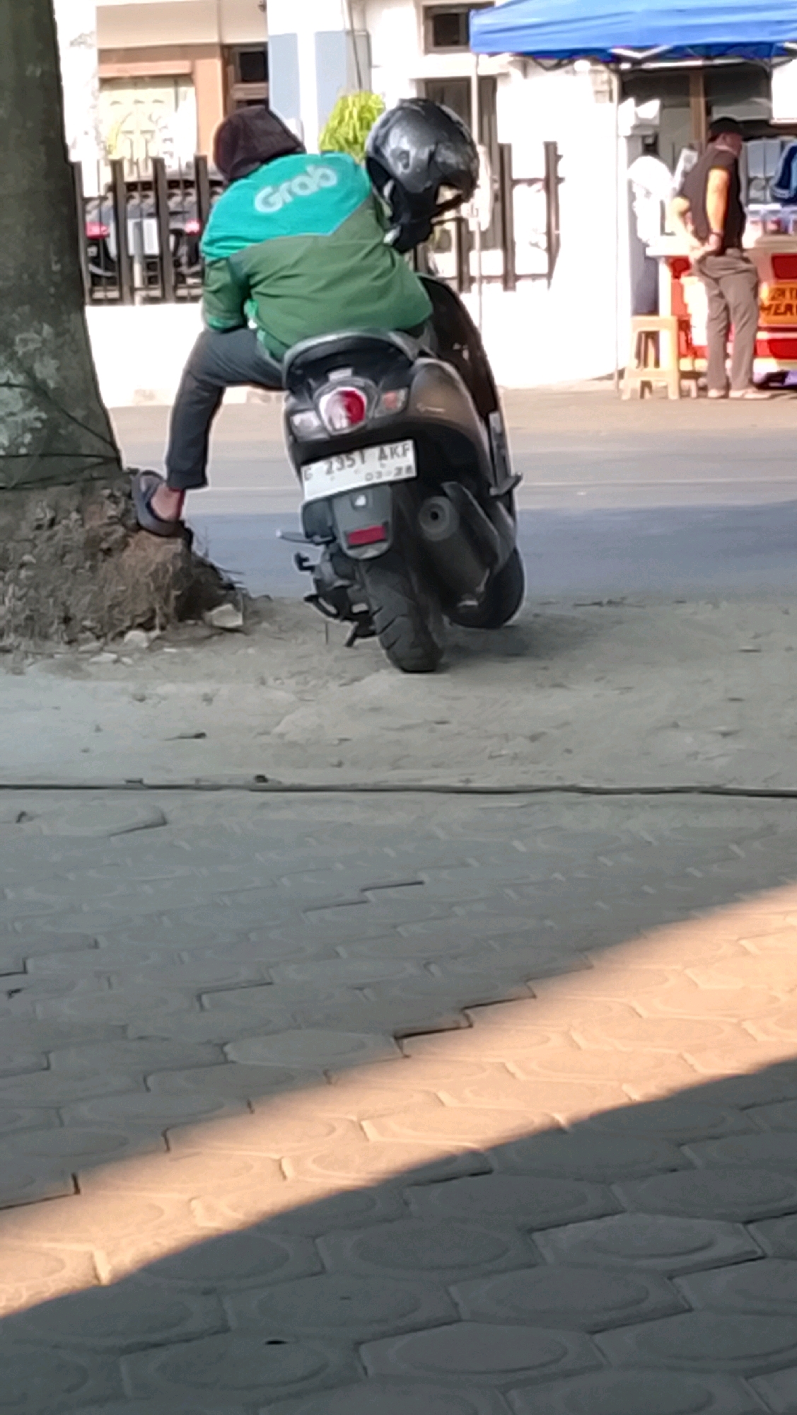 Two individuals, likely a driver and a passenger, are seated on a dark-colored scooter. The passenger, wearing a teal and green jacket with "Grab" logo, has a black helmet on their head. They are positioned behind the driver, who is wearing dark pants and flip-flops. The scooter's license plate, visible at the rear, reads "G 2351 AKF". The scene takes place on a paved sidewalk, adjacent to a dirt embankment with a tree. In the background, a blue awning shelters a food stall, where another person in dark pants and a t-shirt is visible. The overall setting suggests a street scene in the city of Slawi, Indonesia.