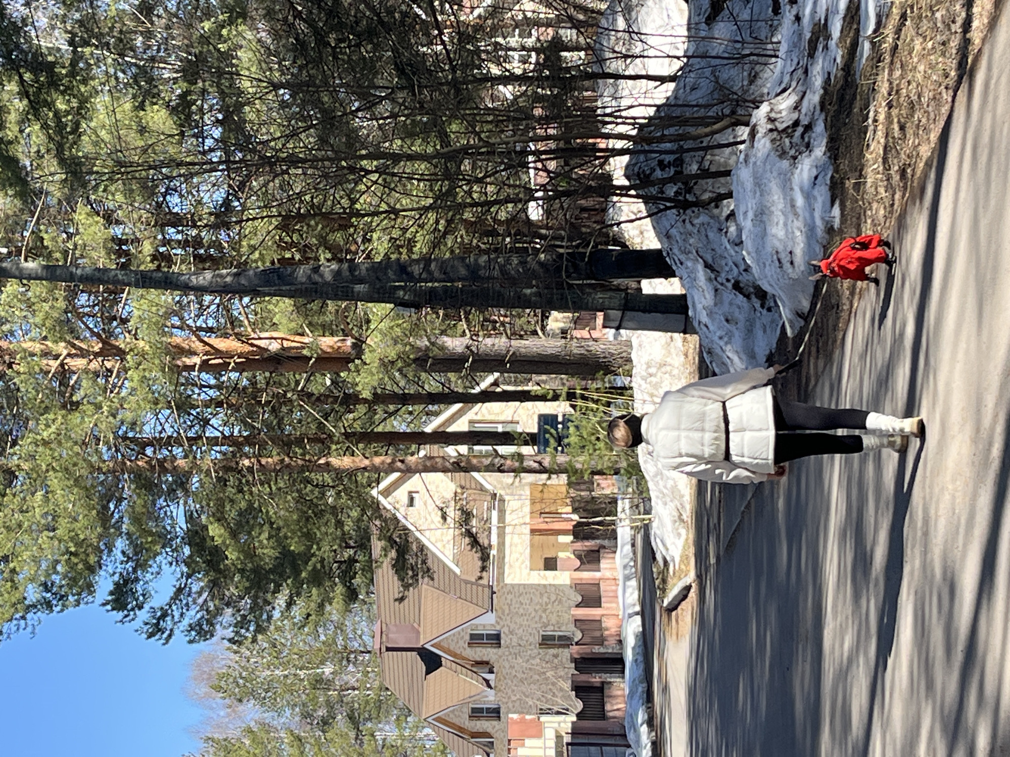 A woman in a white quilted jacket, black leggings, and light-colored boots walks on a paved path. To the left of the path is a snowbank. A person in a red outfit is walking on the path ahead. A building is visible behind the trees to the left. The scene appears to be in Chaikovsky, Russia.