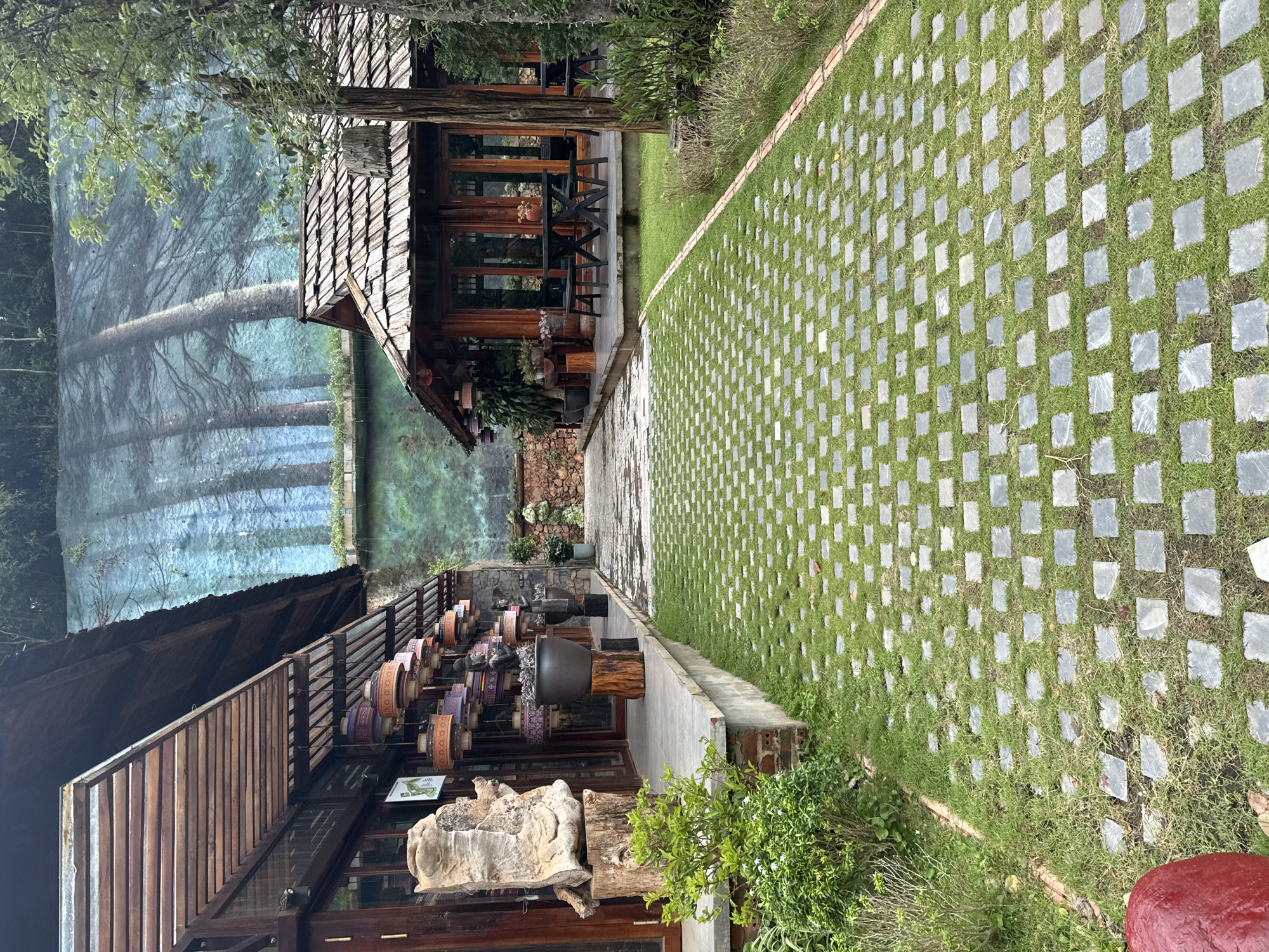 The image depicts an outdoor scene with architectural elements and natural landscaping. On the left, a building with a wooden roof and exterior is visible. Hanging from the eaves are numerous decorative lanterns of various colors and designs. In front of this building, there is a paved area with a large, abstract wooden sculpture on a wooden base. A green bush with small white flowers is growing in the foreground.
To the right, a smaller wooden building with a shingled roof is situated. This building has large glass doors and windows, and outdoor seating with tables and chairs is arranged on its patio.
A pathway composed of green grass interspersed with square grey paving stones leads away from the viewer, towards the buildings. The background features a large mural painted on a wall, depicting a stylized forest scene with tall, slender trees. The overall setting suggests a tranquil, possibly resort or cafe environment. The scene is located in Moc Chau, Vietnam. There are no people visible in the image.