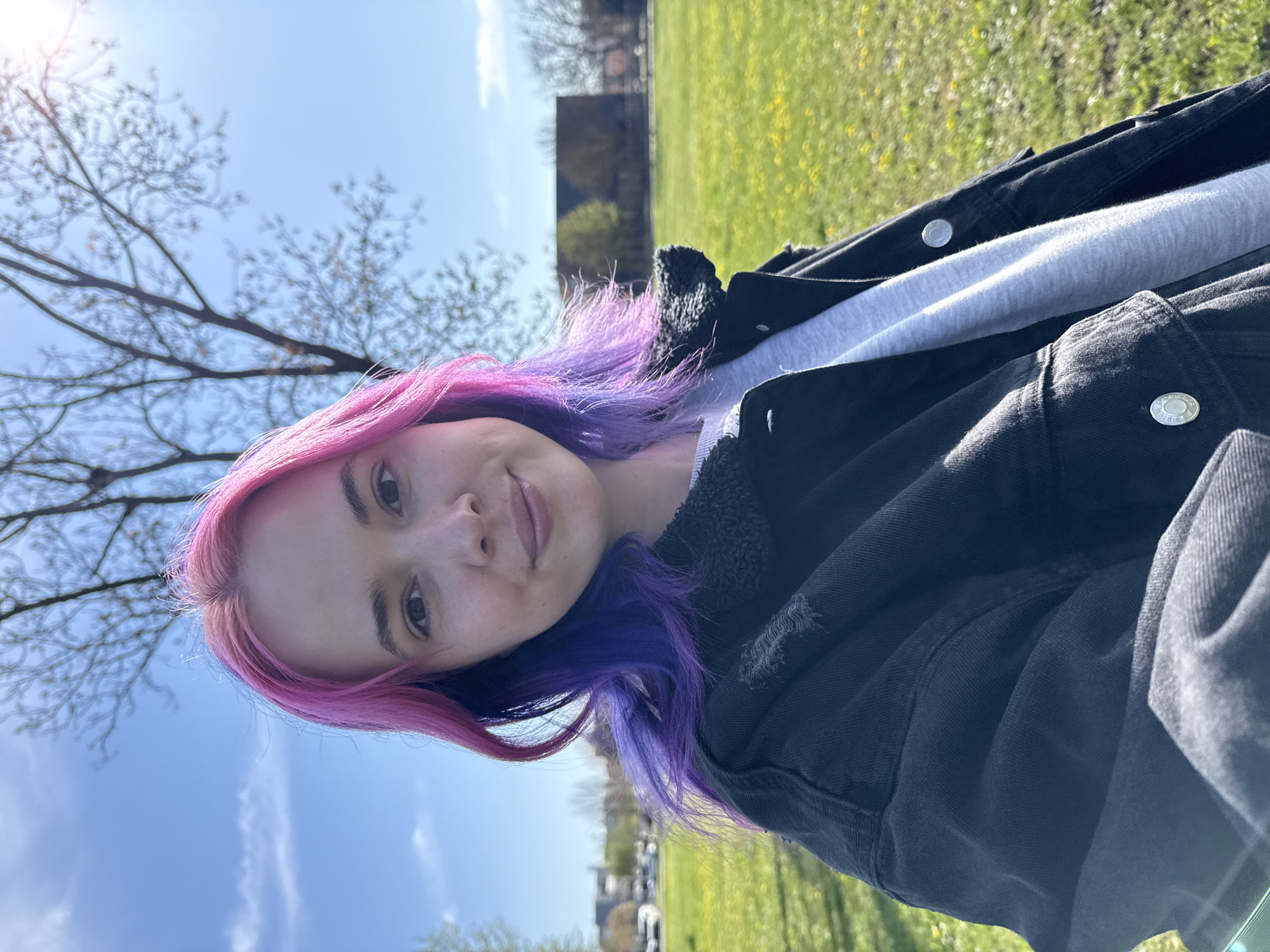 A young woman with pink and purple hair stands outdoors on a sunny day. She is wearing a light gray sweatshirt under a dark denim jacket with a black fleece lining. She is facing the camera with a slight smile.
Behind her, there is a green grassy field, some trees, and buildings in the distance. The sky is blue with a few white clouds.
While the location is indicated as City of Zagreb, Croatia, there are no specific details in the image that confirm the exact location.