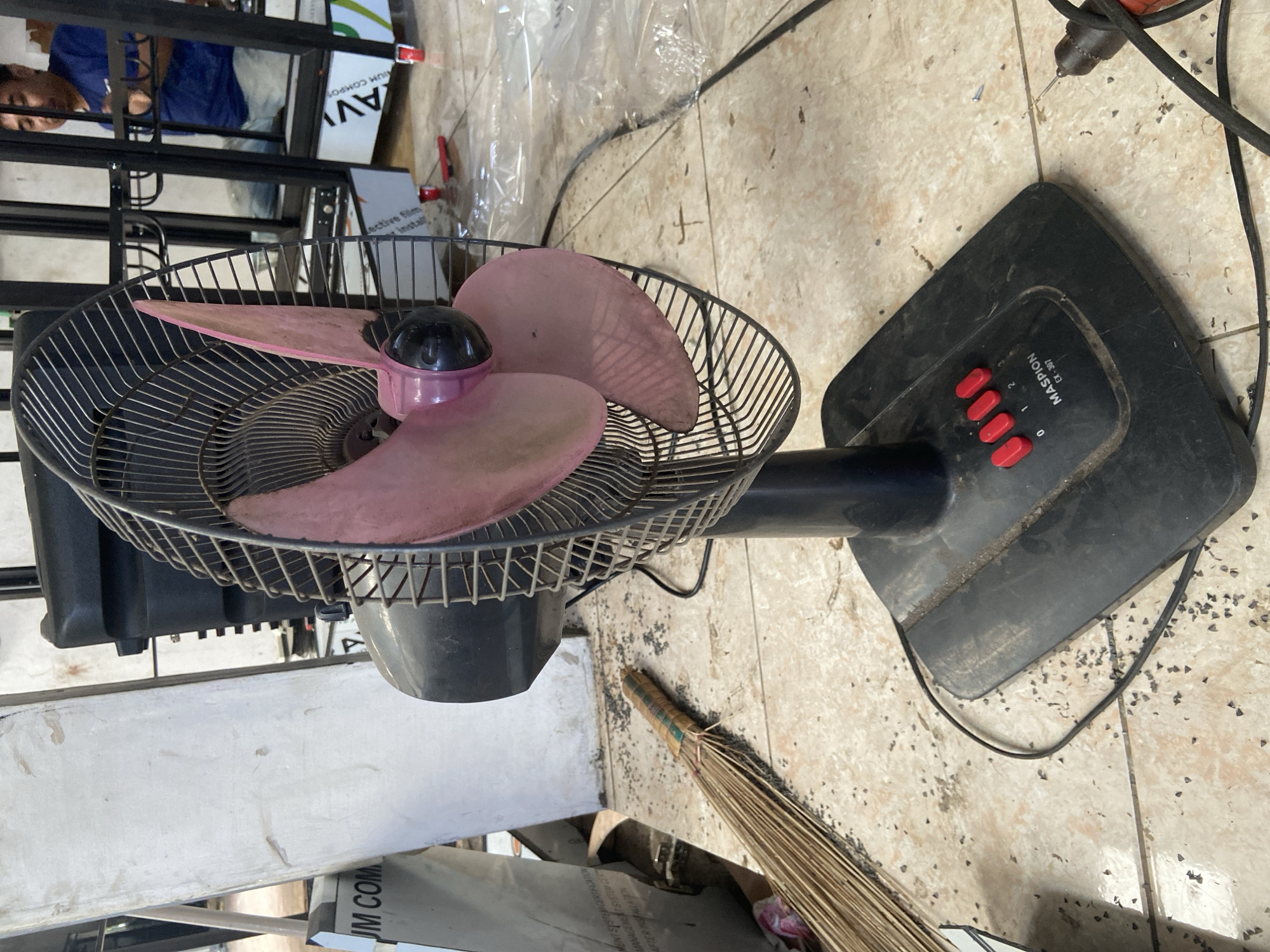 In Plosokandang, Indonesia, a black electric fan with pink blades stands on a tiled floor. The fan's base displays red buttons numbered 0, 1, and 2, with "MASPION CX-307" printed below. To the left of the fan, a broom made of dried plant material is lying on the floor, bound with colorful tape. The floor is littered with small, dark, triangular debris. In the background, a person wearing a blue shirt and light-colored pants is visible through a black metal structure. To the left of the fan, a tall, white, rectangular object with some text is leaning against a surface. A black electrical cord extends from the fan towards the right side of the frame, where it connects to a power source.