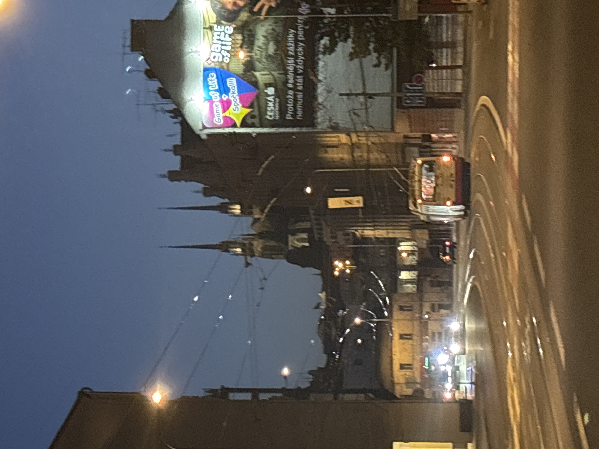 The image depicts an urban street scene at dusk or night. In the foreground, a tram is turning on a curved street. The tram is yellow and red, with its taillights illuminated. The street is paved, with visible lane markings and reflections on the wet surface. In the background, there are several buildings, including a tall, ornate structure with spires, possibly a church or cathedral, illuminated by lights. To the right, a large billboard is visible, advertising "Game of Life" with text in Czech. The billboard features colorful graphics and images. Other buildings in the background appear to be residential or commercial, with illuminated windows. Streetlights are also visible, casting a warm glow on the scene. The sky is a deep blue, indicating the late hour.