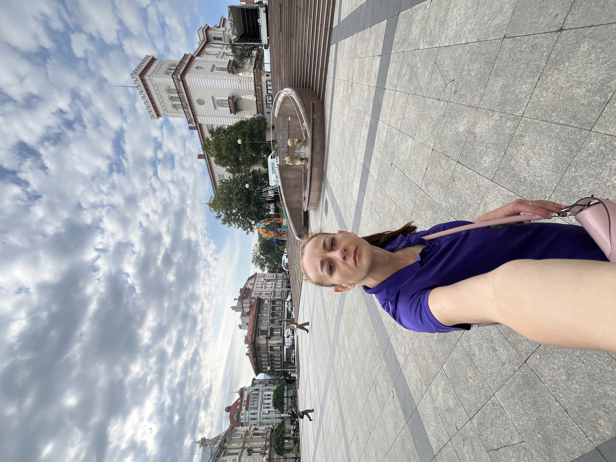 A woman is taking a selfie in a public square. She is wearing a purple shirt and carrying a pink purse. In the background, there are several buildings, a fountain with sculptures, and people walking. The sky is cloudy.