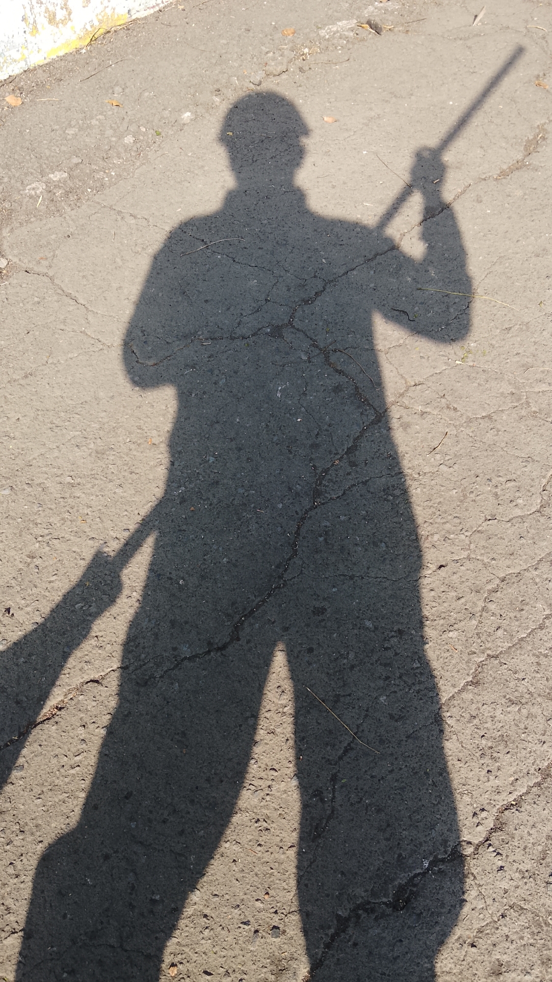 The image shows a long shadow of a person holding a long object, likely a broom, on a cracked asphalt surface. The shadow is cast by sunlight, creating a strong contrast against the lighter ground. The person appears to be standing, with the shadow's head visible at the top and the legs and the object extending downwards and to the left. The environment is outdoors, on what appears to be a street or pathway, with some debris and cracks in the pavement. The location, Tlogobendung, Indonesia, suggests a potentially urban or semi-urban setting. The shadow itself suggests a solitary action of sweeping or holding an implement.