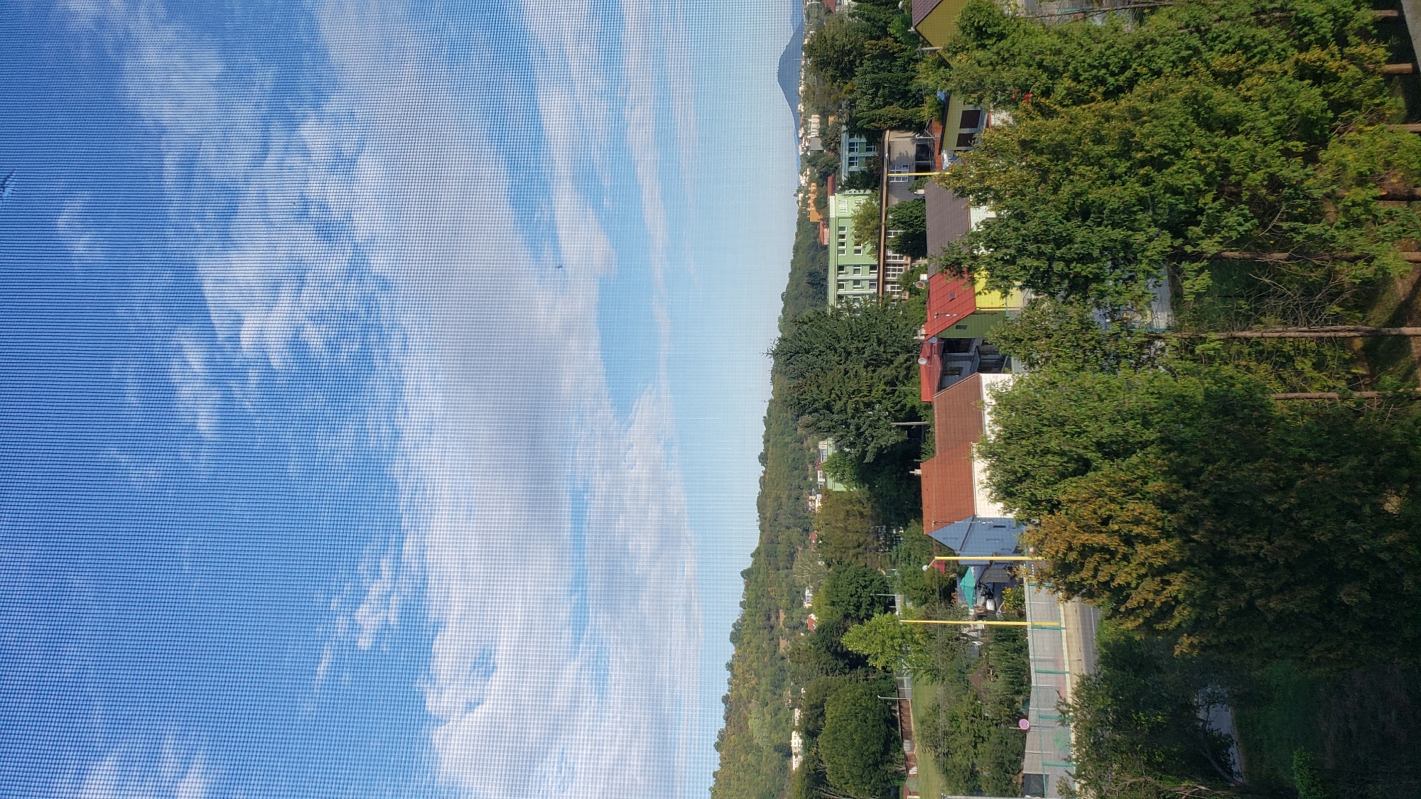 The image shows a view of the city of Prešov, Slovakia, from a slightly elevated position. The foreground is dominated by various types of trees, including tall, slender evergreens and deciduous trees with autumn foliage. Behind the trees, several houses with red-tiled roofs are visible, along with some residential buildings in the background. A green building with multiple windows stands out among the other structures. The landscape behind the city rises into rolling hills covered with trees. The sky is blue with scattered white clouds. A mesh screen is superimposed on the image, indicating it was taken through a window. The scene appears to be a residential area on a clear day.