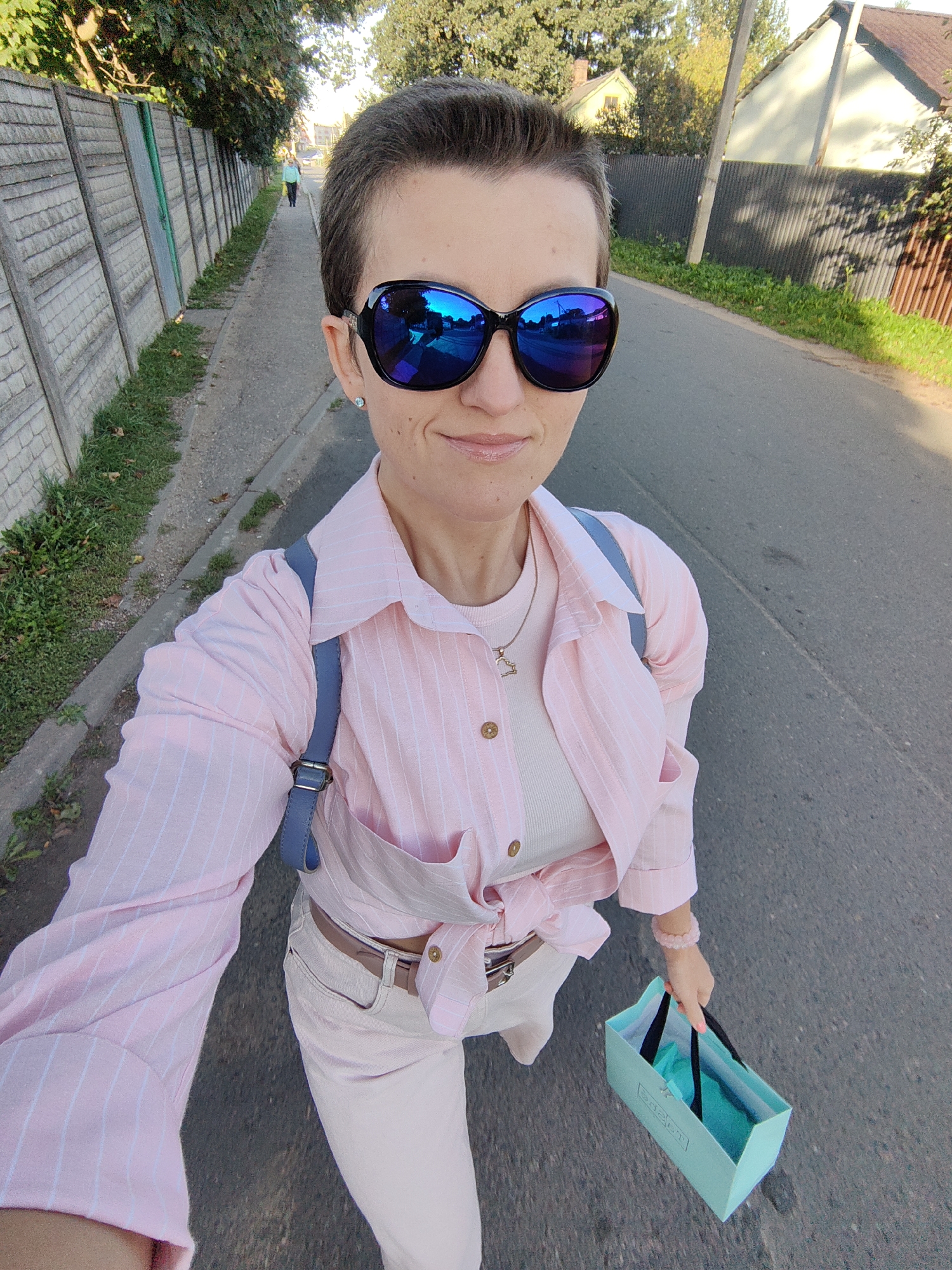 A woman with short, dark hair and sunglasses is walking on a paved road in Fanipal, Belarus. She is wearing a pink striped shirt, a white tank top, and light-colored pants. She is carrying a light blue shopping bag in her right hand and has a light blue strap across her chest, possibly from a backpack or purse. The road is lined with a concrete fence and grass on the left, and houses and trees on the right. The sun is shining, casting shadows on the road and houses. In the background, another person is walking away from the camera on a sidewalk.