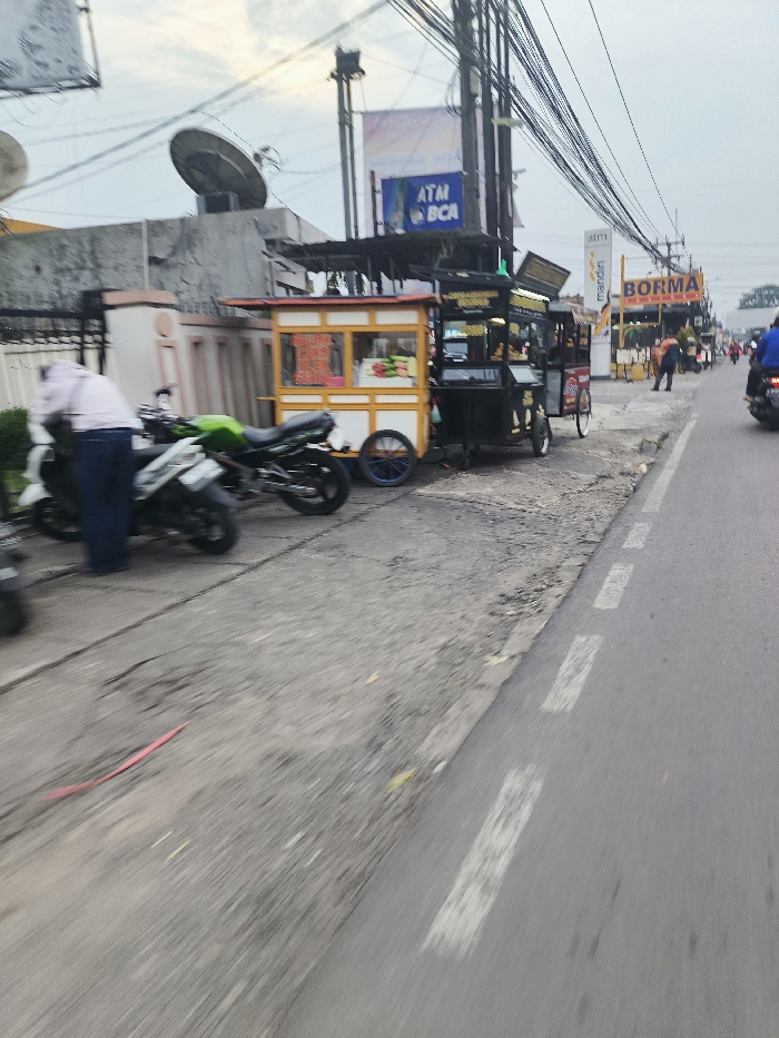 The image shows a street scene in Cileunyi, Indonesia, at coordinates -6.937 latitude and 107.732 longitude. A man in a light pink shirt and blue pants stands next to a green and black motorcycle. The motorcycle is parked on the side of the road, facing a street vendor selling food from a black and yellow cart. The cart is parked on the sidewalk along the road, which has a white lane divider. The vendor’s cart is situated near a sign that reads "Borma" in white letters on a blue background. Another person in a black shirt is visible walking away from the camera on the sidewalk. The road is paved and rough. The scene is partially obscured by a blurred background.