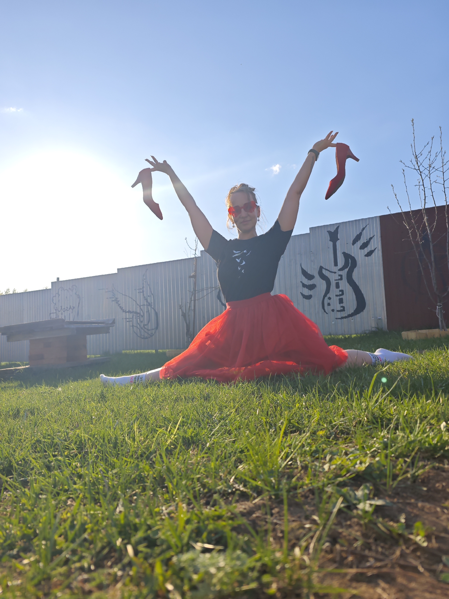 A woman is performing a split on a lawn in Markovo, Russia. She is wearing a black t-shirt, a red tutu, white socks, and red heart-shaped sunglasses, and she is holding a red high-heeled shoe in each hand, arms raised. The background includes a grey fence with a black guitar painted on it, a wooden structure, and a clear, blue sky. The setting is outdoors with green grass covering the ground.