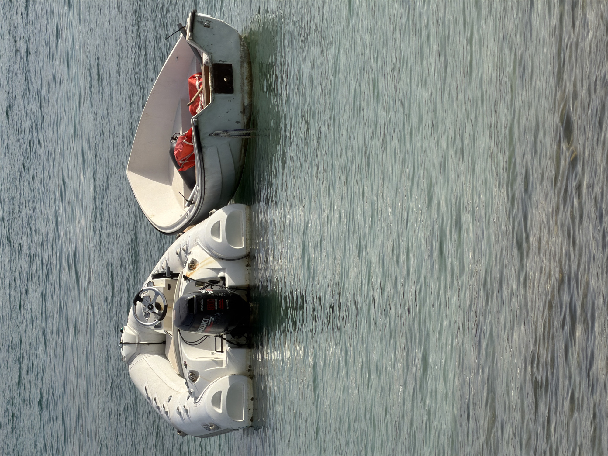 Two small boats are moored on the calm, clear turquoise waters of the Mediterranean Sea, likely near the coast of Calvià, Spain. The boat on the left is a white inflatable dinghy with an outboard motor and a steering wheel. The boat on the right is a small, white, rigid-hulled boat with two orange life vests inside and a wooden paddle. The water is slightly rippled, reflecting the bright daylight. The background is out of focus, showing only the water and a sliver of what might be the shoreline in the distance.