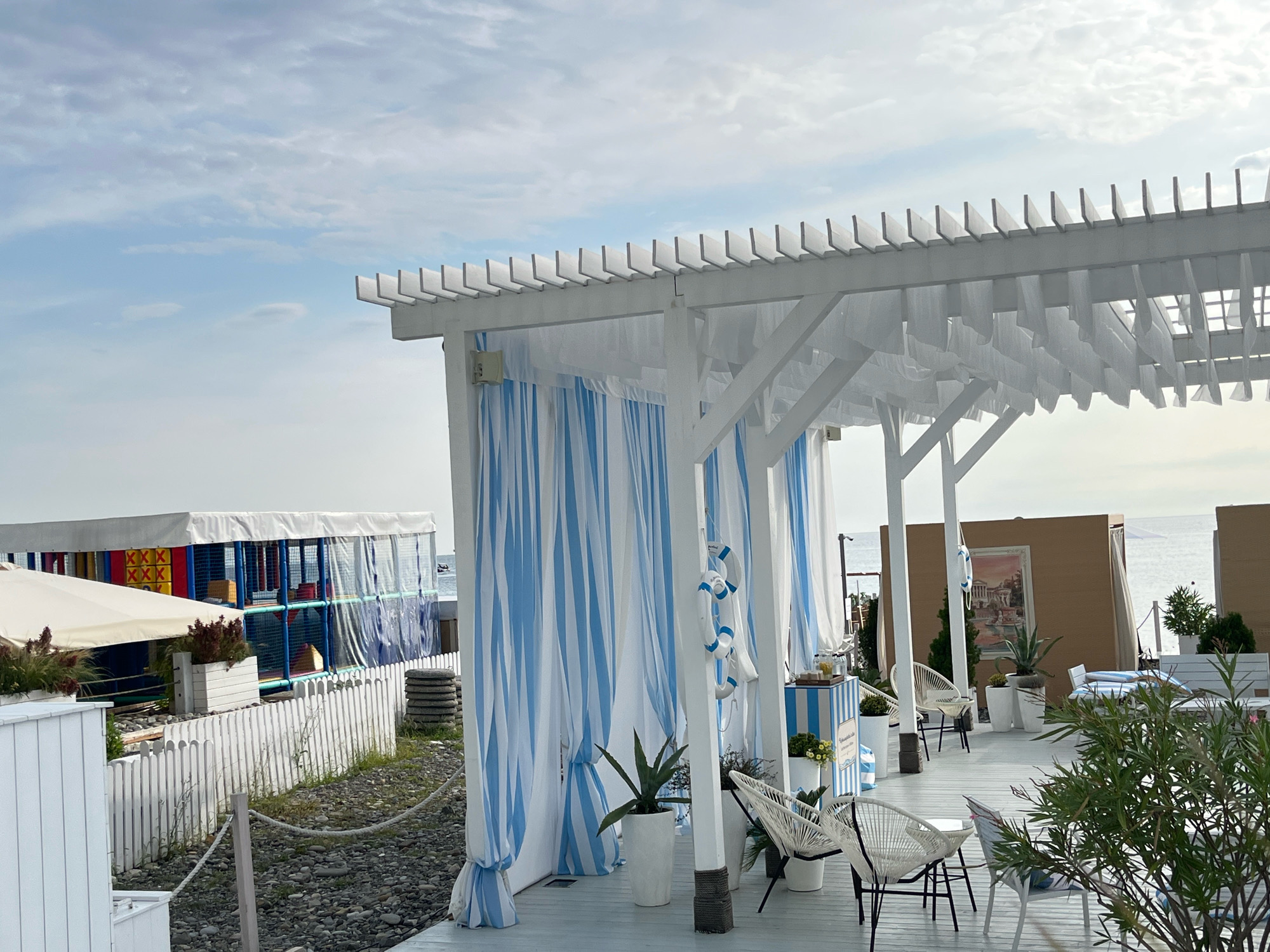 The image depicts an outdoor seating area with white wooden structures and blue and white striped curtains. The scene is located in Sochi, Russia, a coastal city known for its beaches and resort areas. In the background, there is a colorful, enclosed play area, likely for children, covered with a white tarp. A white picket fence separates the seating area from a gravel path. The sea is visible in the distance. The seating area features white chairs with woven seats and black frames, as well as potted plants. Two white life preservers are hanging from a post. The overall impression is a relaxed, beachside atmosphere.