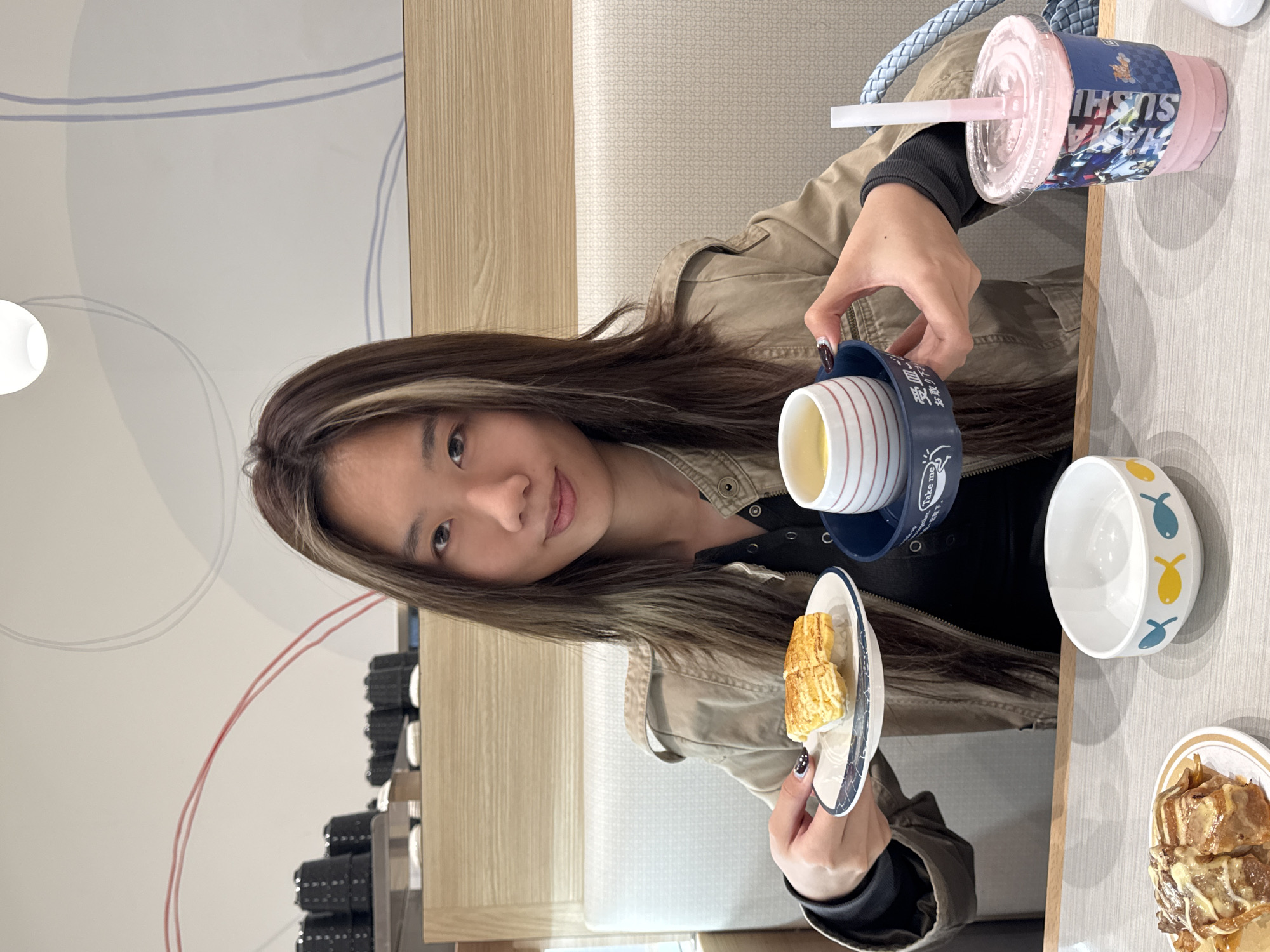 A young woman with highlighted brown hair is seated at a table in a restaurant. She holds a small plate with a piece of food. On the table, there is a cup of tea, a bowl with fish designs, a pink drink in a plastic cup, and a plate of food with a dark sauce. The background shows a restaurant interior with a wall featuring curved lines and a wooden panel. Based on the "Hana Sushi" logo visible on the pink drink, it's likely a Hana Sushi branch in Nan'an District, China.