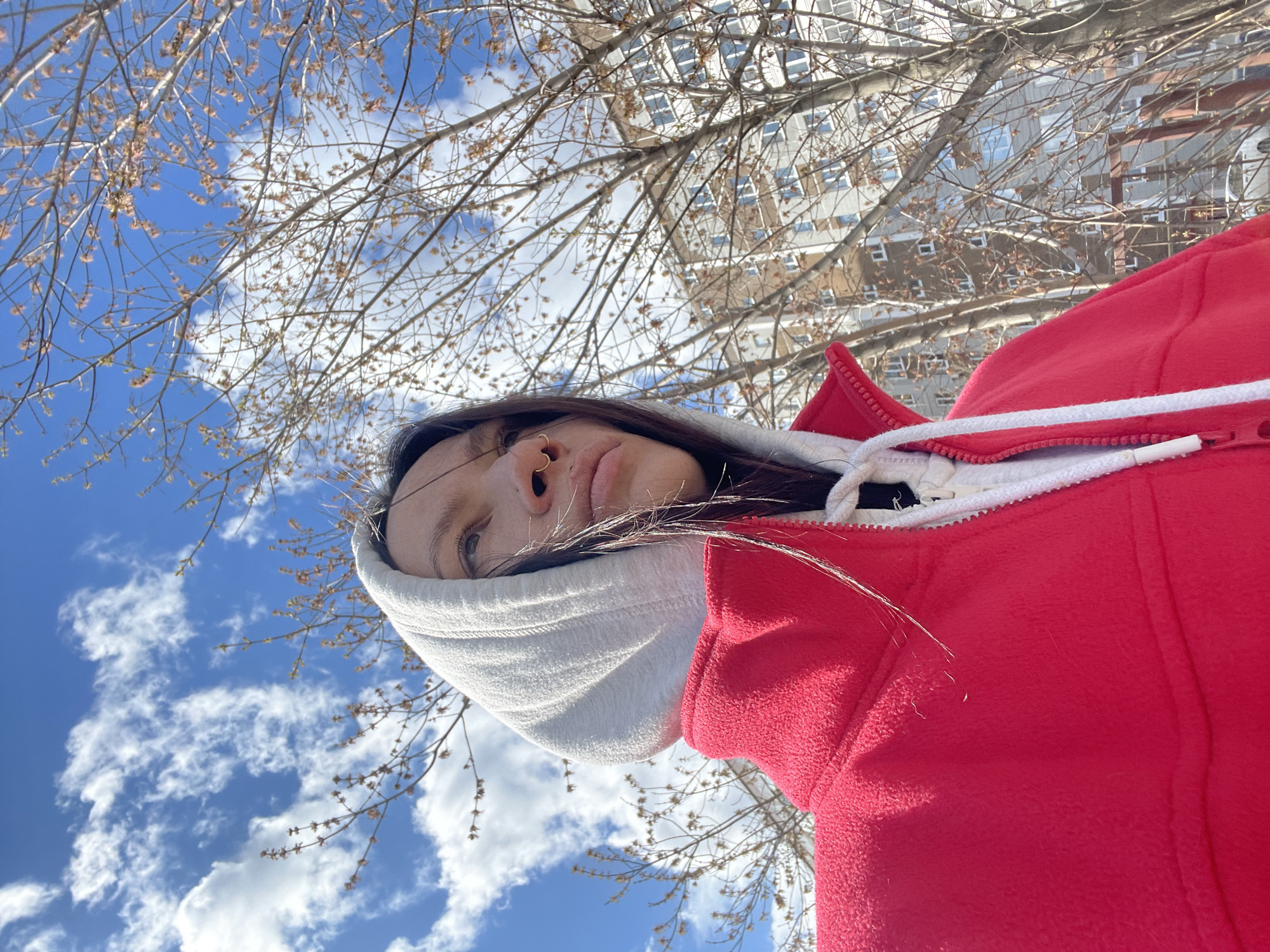 A low-angle shot captures a person looking upwards, framed against a vibrant blue sky filled with white, fluffy clouds. The person, visible from the chin to the top of the head, appears to be a woman with dark hair and a gold nose ring on her right nostril. She is wearing a bright red fleece jacket with a white hooded collar and a white drawstring.
Numerous bare tree branches, adorned with small buds, crisscross the upper and middle sections of the image, suggesting early spring or late winter. Through these branches, parts of a tall, multi-story building with many windows can be seen, indicating an urban setting. The bright, clear daylight suggests the photograph was taken during the day, likely midday. The scene conveys a calm, outdoor moment in Izhevsk, Russia. There is no visible text in the image.
