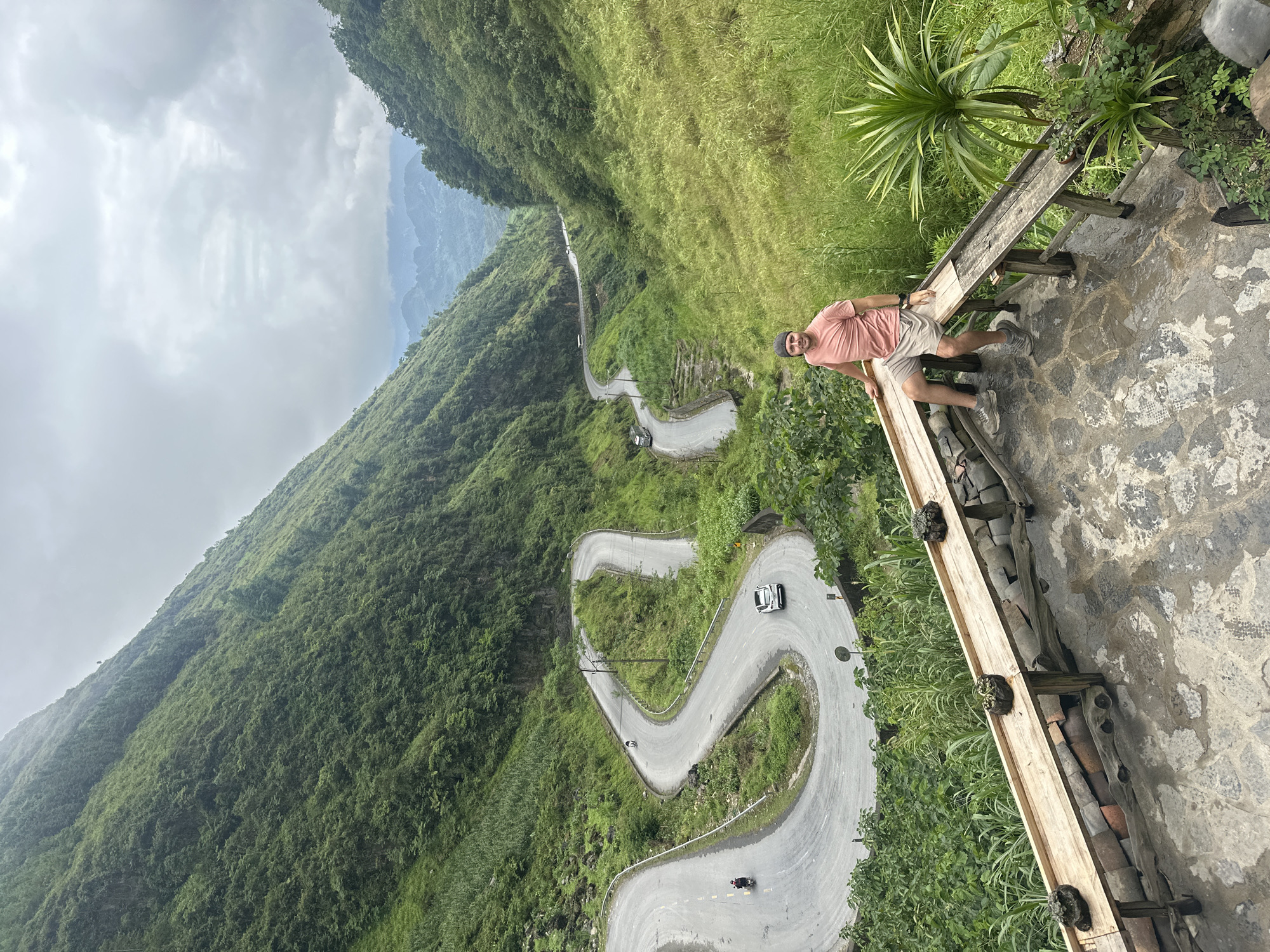 A man stands on a paved overlook next to a wooden railing, gazing toward the camera. He wears a pink t-shirt, khaki shorts, and a baseball cap. Below, a winding road with several hairpin turns traverses a steep, lush green hillside. A white car and a motorcycle are visible on the road. The sky is overcast. The location is Phố Bảng Commune, Vietnam.