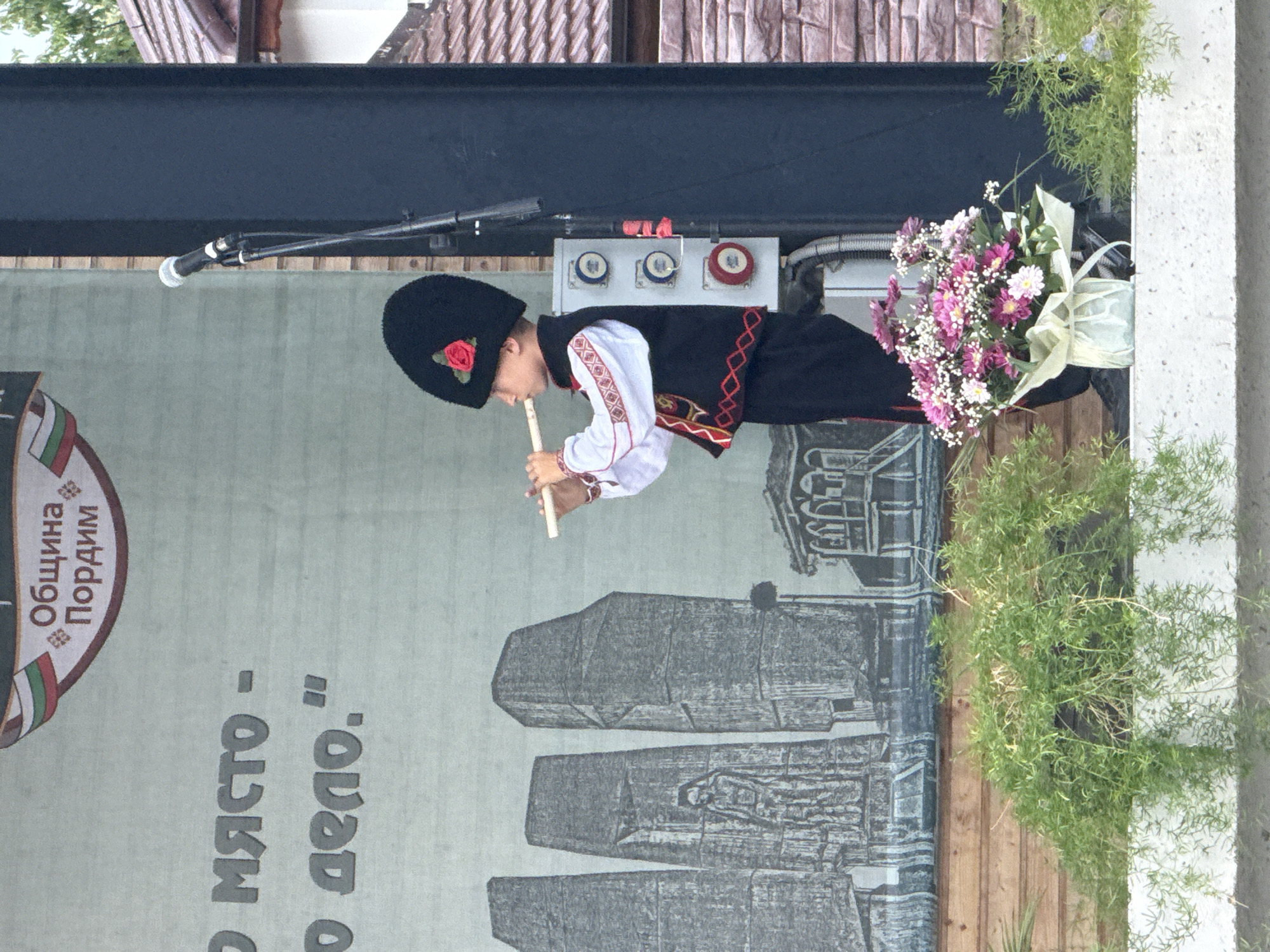 A young boy in traditional Bulgarian attire, including a tall black hat with a red rose, is playing a wooden flute on a stage. He wears a white embroidered shirt, a dark vest with red trim, and dark pants. The backdrop is a large banner featuring a grey, stylized image of monumental structures and possibly historical figures, along with Bulgarian text. To the right of the boy is a black speaker and a microphone stand. In front of the stage, there is lush green foliage, and on the edge of the stage, a vase of pink and white flowers. The scene appears to be an outdoor event in the town of Pordim, Bulgaria, likely a cultural performance.