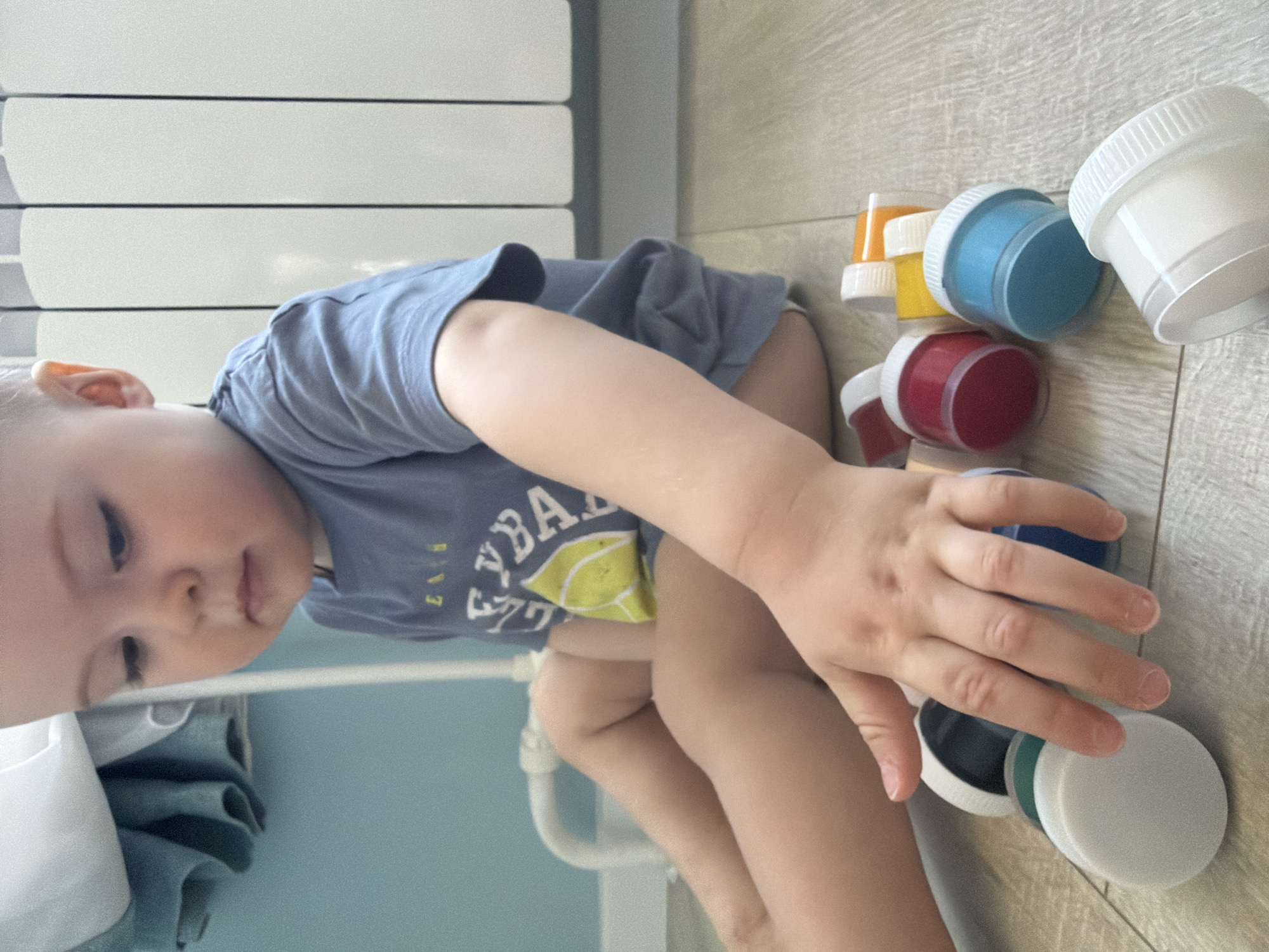 A young child is sitting on a light-colored, wood-patterned floor next to an array of open paint containers. The paint containers are in various colors, including white, yellow, blue, red, and black. The child is reaching towards the paint with their right hand. The child is wearing a light blue shirt with a graphic design on the front. To the left, there is a white radiator with a light blue wall in the background. The location is presumably inside a home. It is not possible to determine the location of Votkinsk, Russia from the provided image.