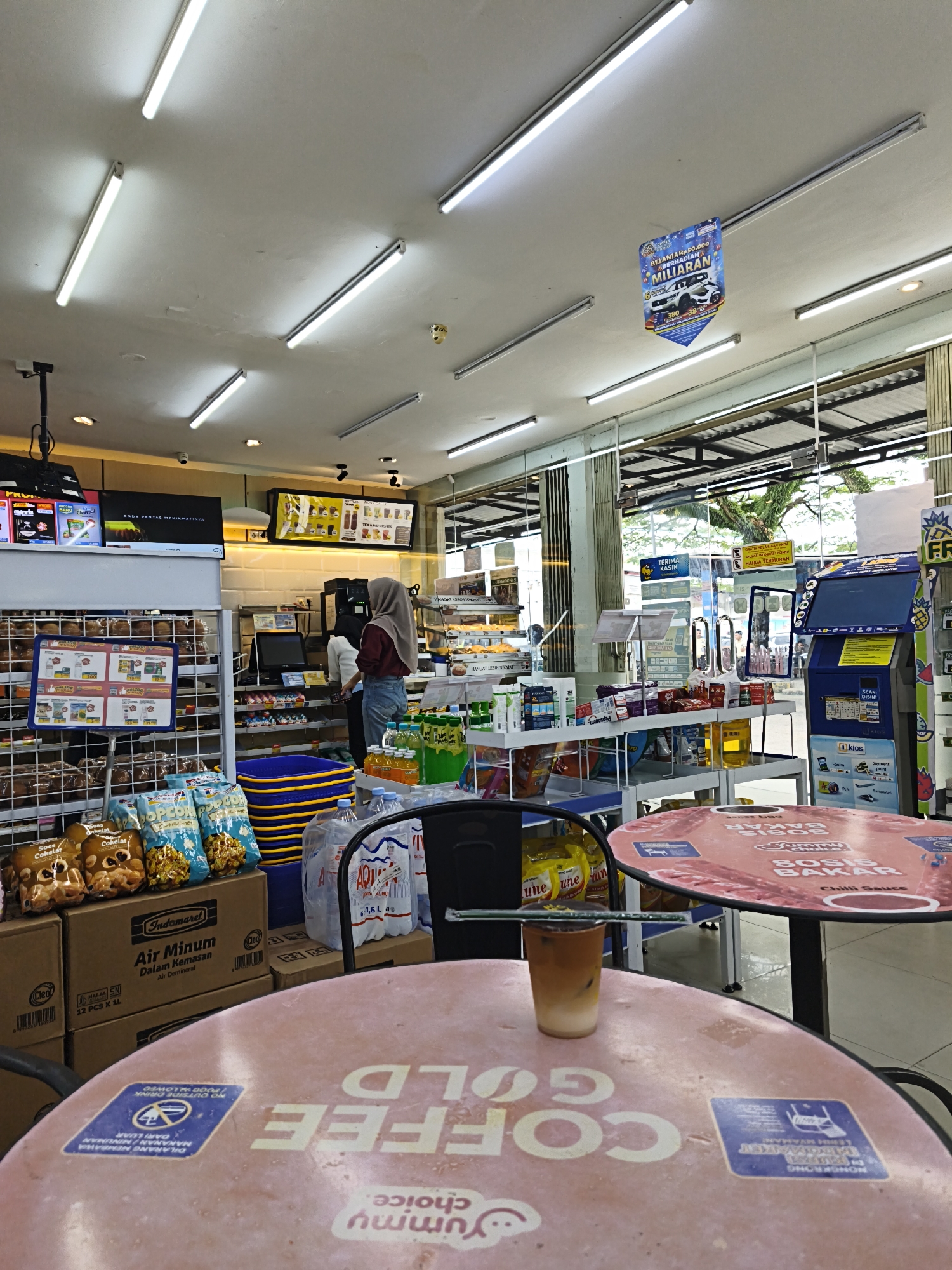 The image depicts the interior of an Indomaret convenience store in Bandar Lampung, Indonesia. The store is well-lit by fluorescent ceiling lights and features rows of shelves stocked with various products. In the foreground, two pink circular tables are visible, one with a plastic cup containing a beverage and a straw. The table closer to the viewer has text and stickers on its surface, including the words "COFFEE GOLD" and a blue sticker with Indonesian text.
In the middle ground, a cashier counter with a computer monitor and a payment machine is attended by two individuals. Behind the counter, shelves display more products, including bottled drinks and snacks. A large blue stacking basket is also present. To the right, a blue self-service kiosk is positioned near the entrance.
The background shows the store's exterior through large glass windows, revealing trees and the sky. A banner hanging from the ceiling promotes a lucky draw with a car as a prize. The overall impression is that of a typical convenience store environment, with merchandise neatly displayed and customers or staff present.