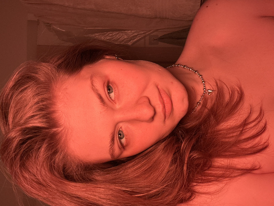 An indoor scene in the city of Noviy, Russia, illuminated by a dominant red light source. The primary subject is an individual, identified as female, positioned centrally, with her head and upper torso visible. She has long, reddish-brown hair draped over her left shoulder and partially across her chest. Her eyes are light blue-green, directed towards the upper right of the frame. Her lips are closed. A silver-colored beaded necklace with a small, silver heart-shaped pendant is worn around her neck, resting on her bare skin. The background is indistinct, showing faint vertical lines on the right, consistent with a doorframe or hanging fabric, all bathed in the red light. The individual exhibits a neutral or subtly smiling facial expression.